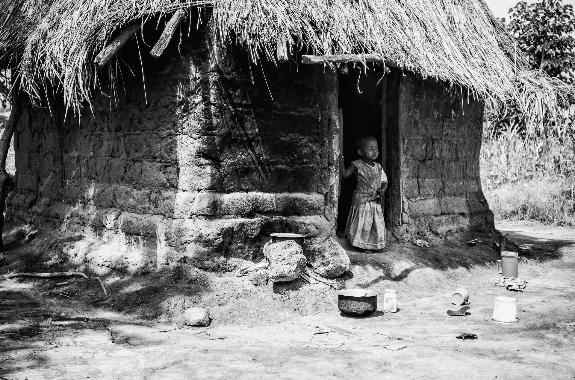 A black and white portrait photograph of a young African child standing in the doorway of a simple mud hut.