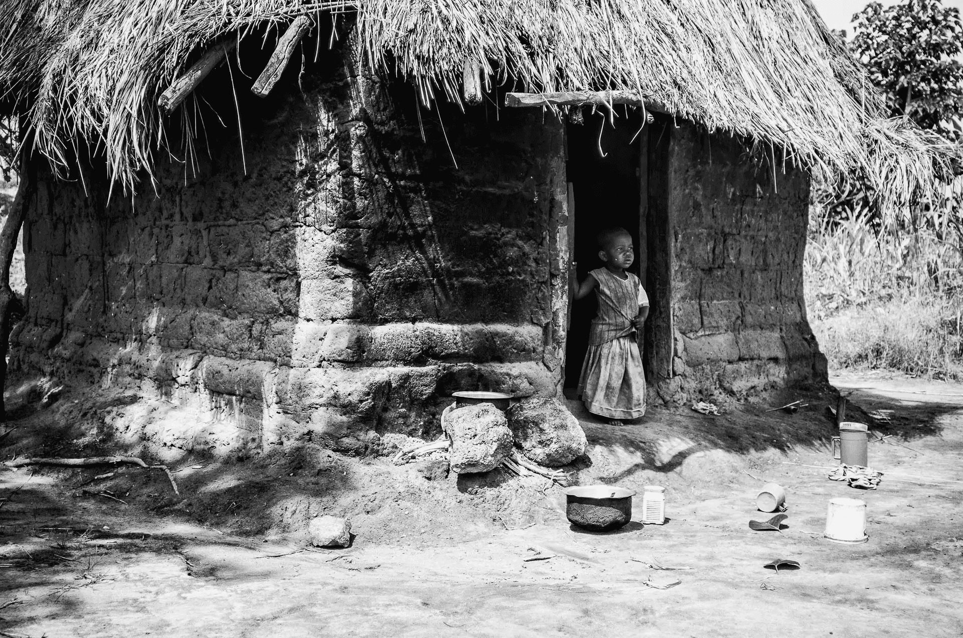 A black and white portrait photograph of a young African child standing in the doorway of a simple mud hut.