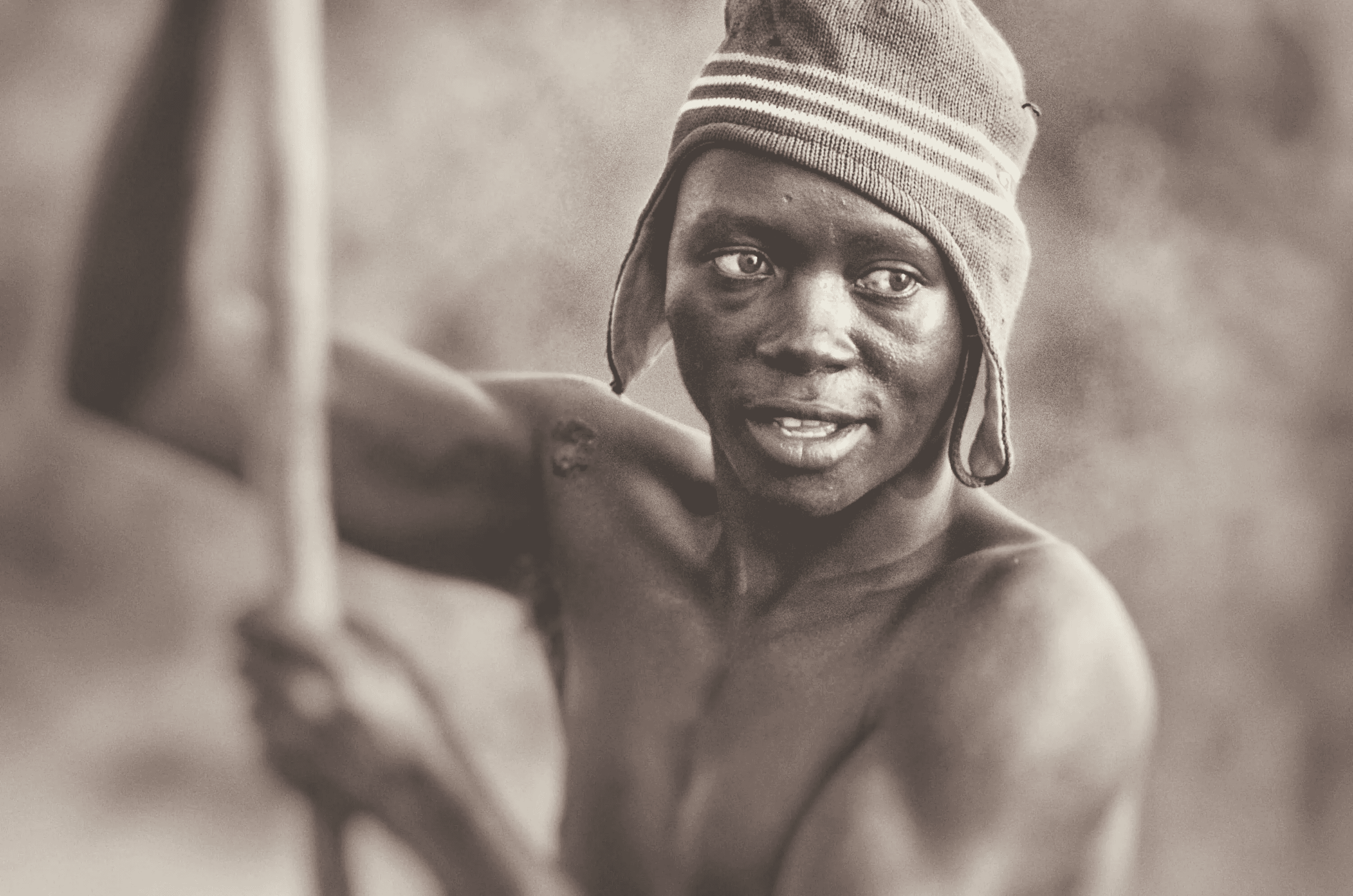 A close-up, sepia-toned portrait photograph of a Ugandan fisherman. He is wearing a knitted hat with stripes and is holding a long wooden pole, possibly a fishing rod or part of a boat.