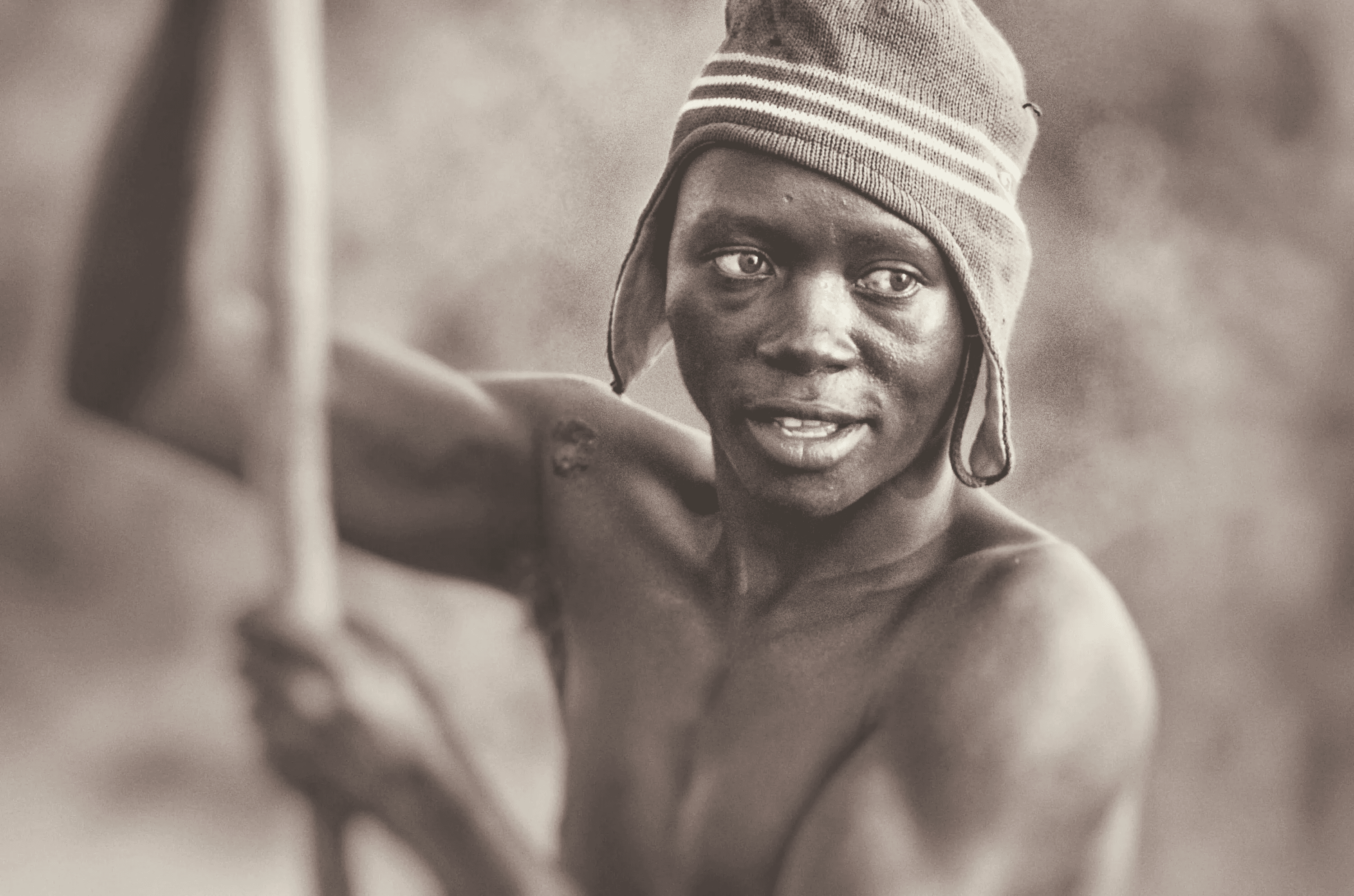 A close-up, sepia-toned portrait photograph of a Ugandan fisherman. He is wearing a knitted hat with stripes and is holding a long wooden pole, possibly a fishing rod or part of a boat.
