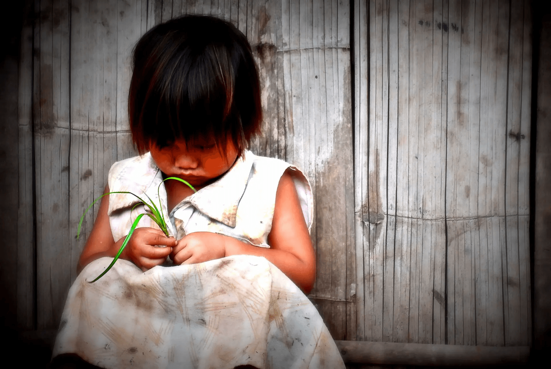 A tender portrait photograph of a young child, with dark hair and a simple white dress, sitting against a textured bamboo wall.