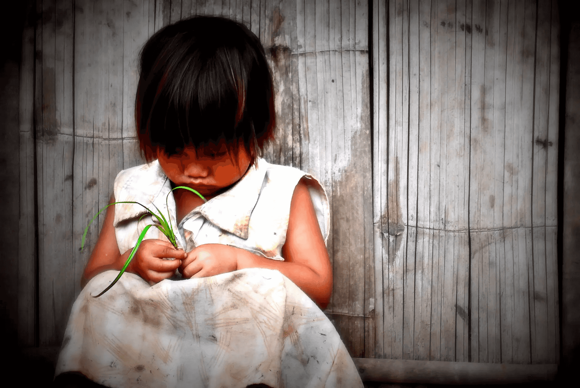 A tender portrait photograph of a young child, with dark hair and a simple white dress, sitting against a textured bamboo wall.