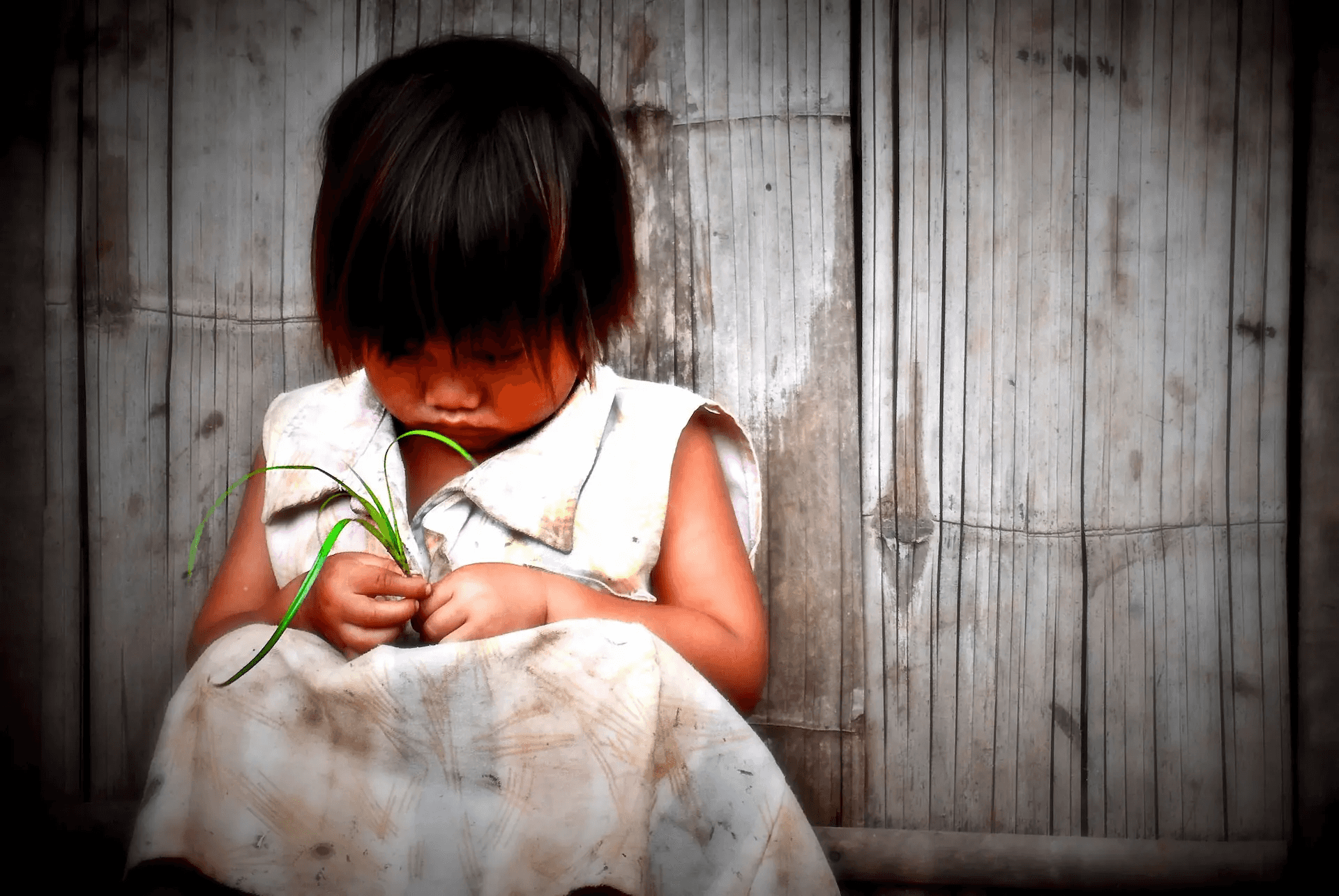 A tender portrait photograph of a young child, with dark hair and a simple white dress, sitting against a textured bamboo wall.