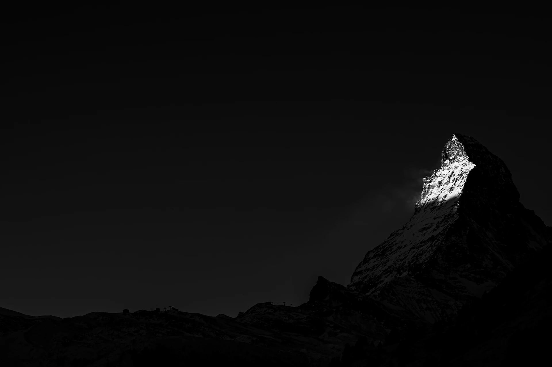 A dramatic black and white photograph of the Matterhorn mountain peak, with the first rays of morning sunlight catching the summit, while the rest of the mountain remains in shadow.