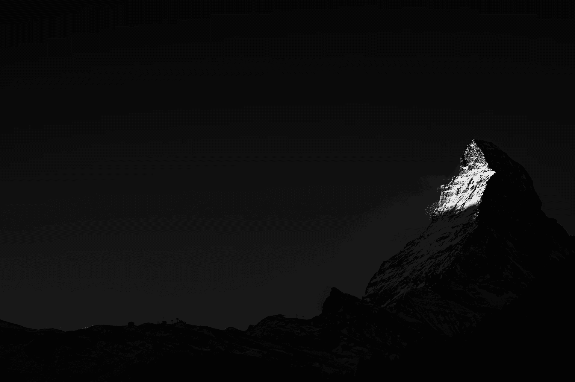 A dramatic black and white photograph of the Matterhorn mountain peak, with the first rays of morning sunlight catching the summit, while the rest of the mountain remains in shadow.