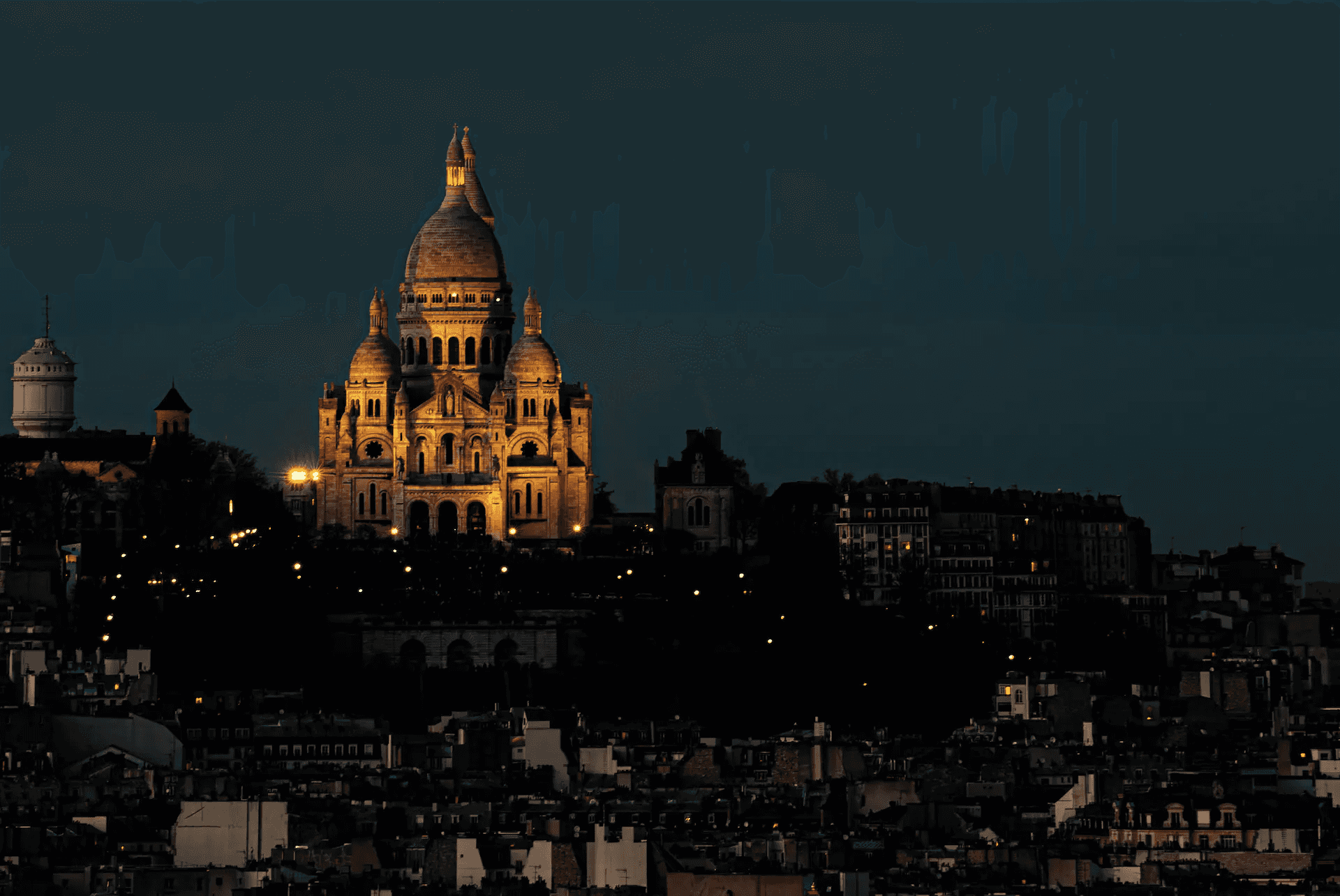 A long shot photograph capturing the Basilica of Sacré-Cœur de Montmartre in Paris, illuminated against a deep blue evening sky.