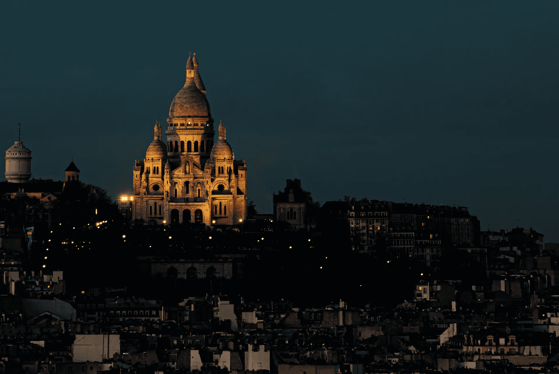 A long shot photograph capturing the Basilica of Sacré-Cœur de Montmartre in Paris, illuminated against a deep blue evening sky.