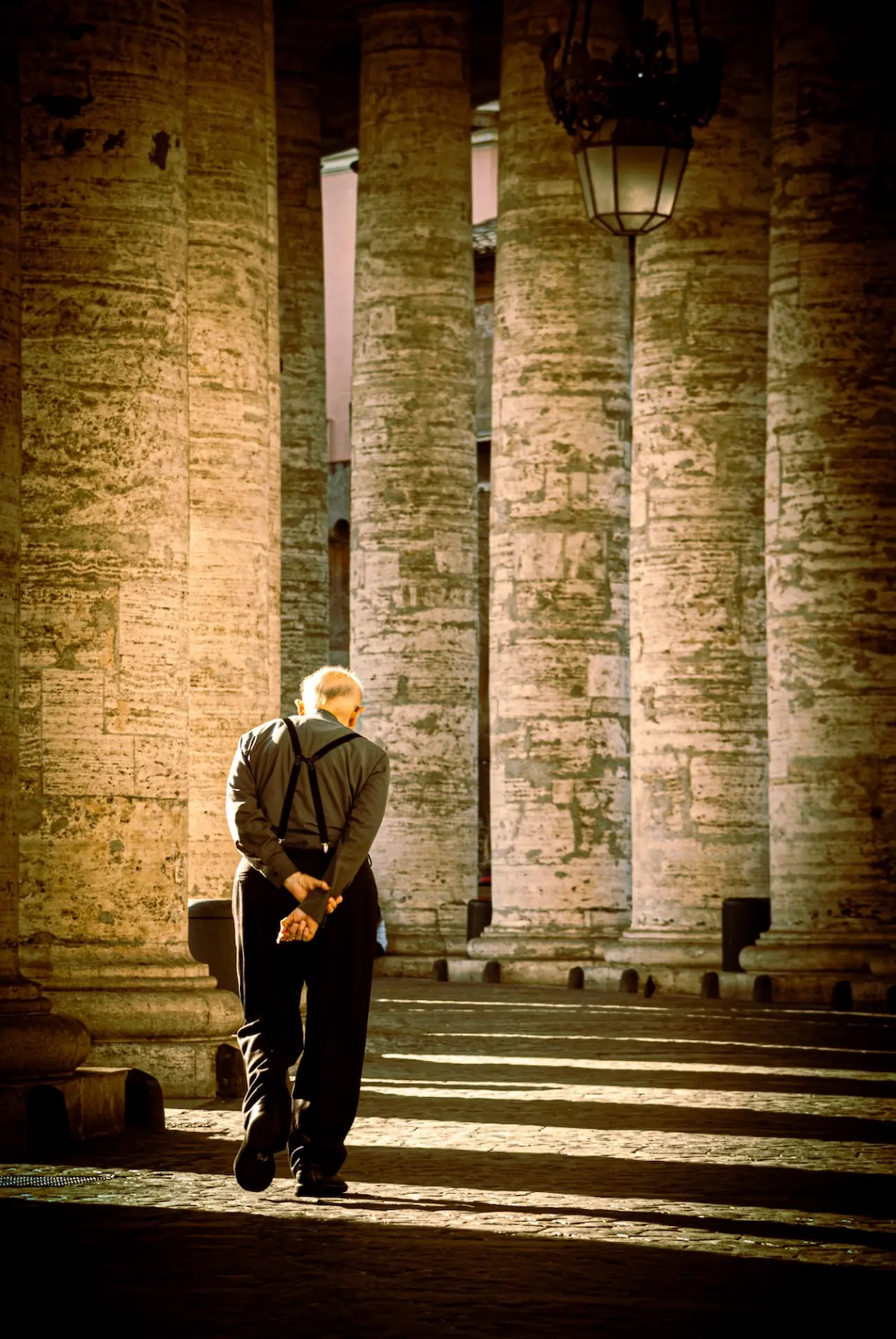 A photograph capturing an elderly priest walking away from the viewer under the grand colonnade of St.