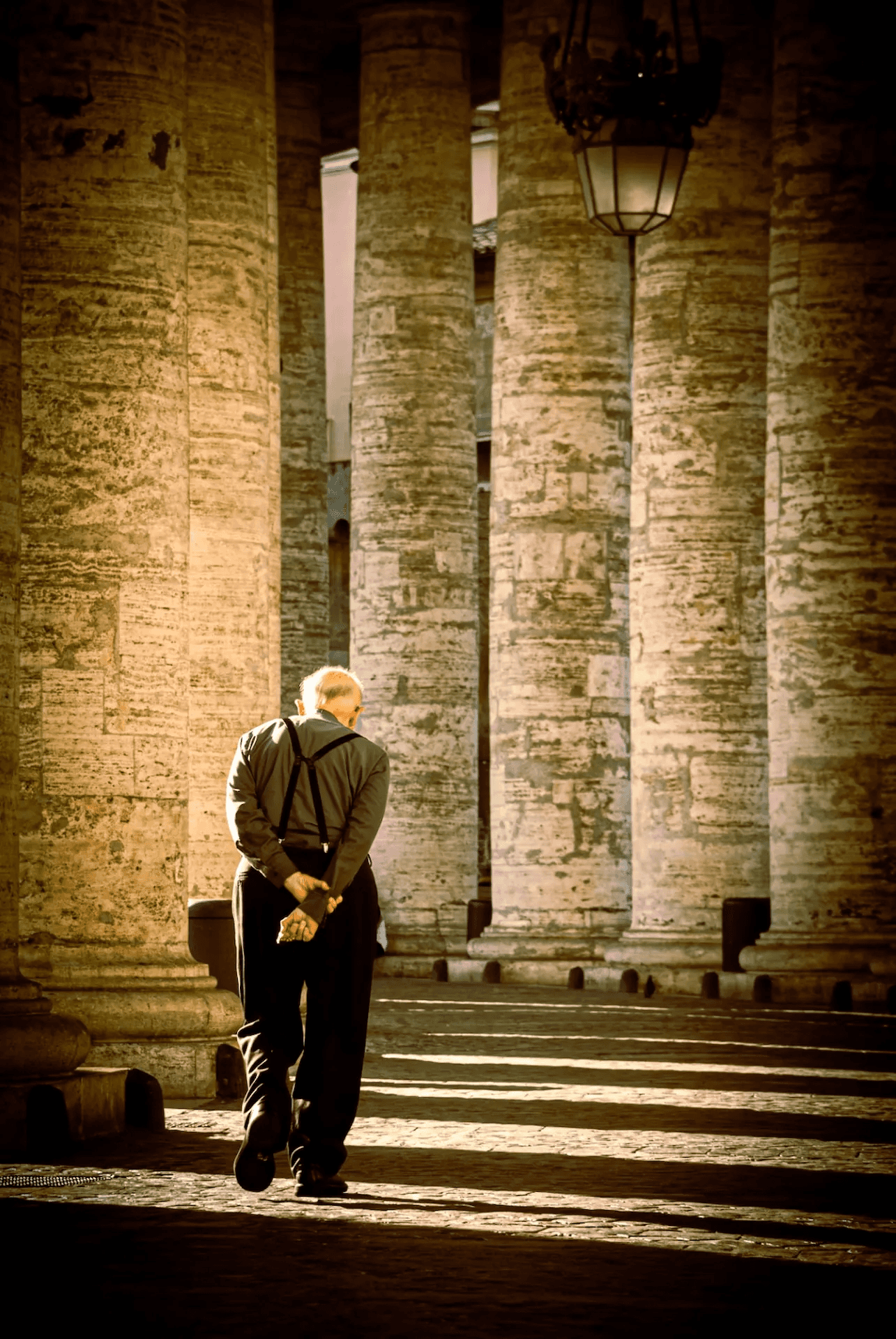 A photograph capturing an elderly priest walking away from the viewer under the grand colonnade of St.