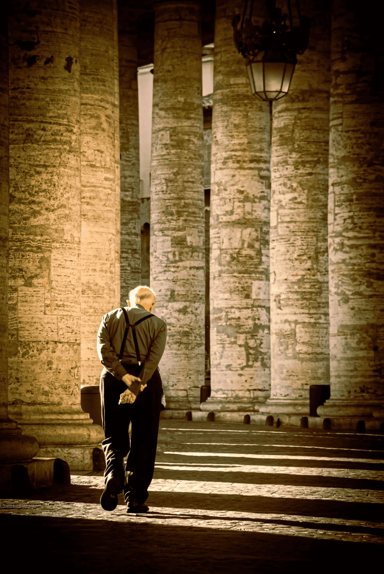 A photograph capturing an elderly priest walking away from the viewer under the grand colonnade of St.
