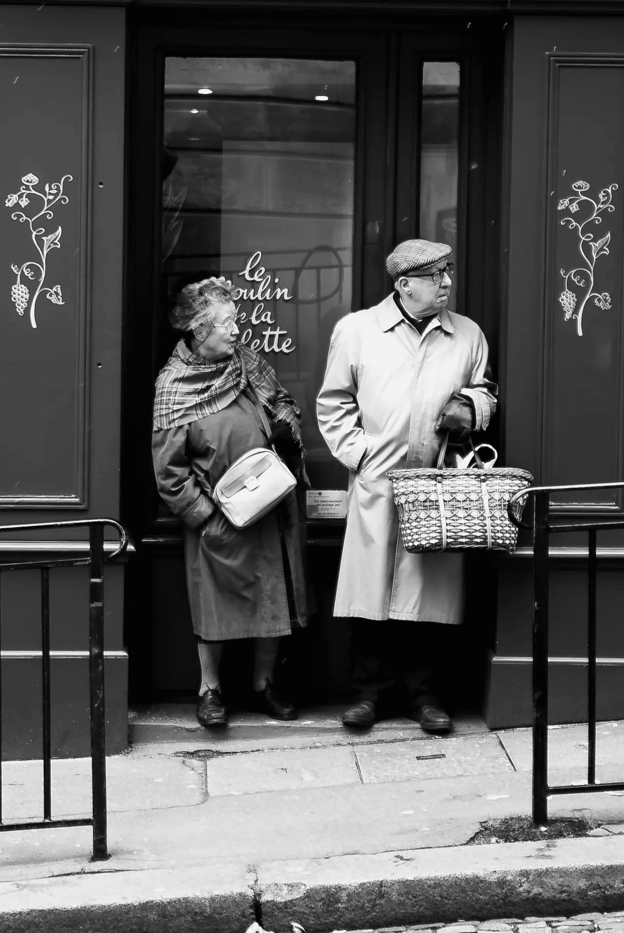 A black and white street photograph of an elderly couple standing in the doorway of a Parisian shop. The woman, on the left, is wearing a coat and scarf and carrying a handbag, looking slightly to the side with a thoughtful expression.