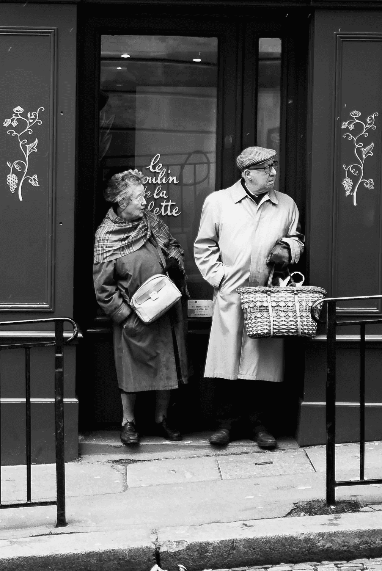 A black and white street photograph of an elderly couple standing in the doorway of a Parisian shop. The woman, on the left, is wearing a coat and scarf and carrying a handbag, looking slightly to the side with a thoughtful expression.