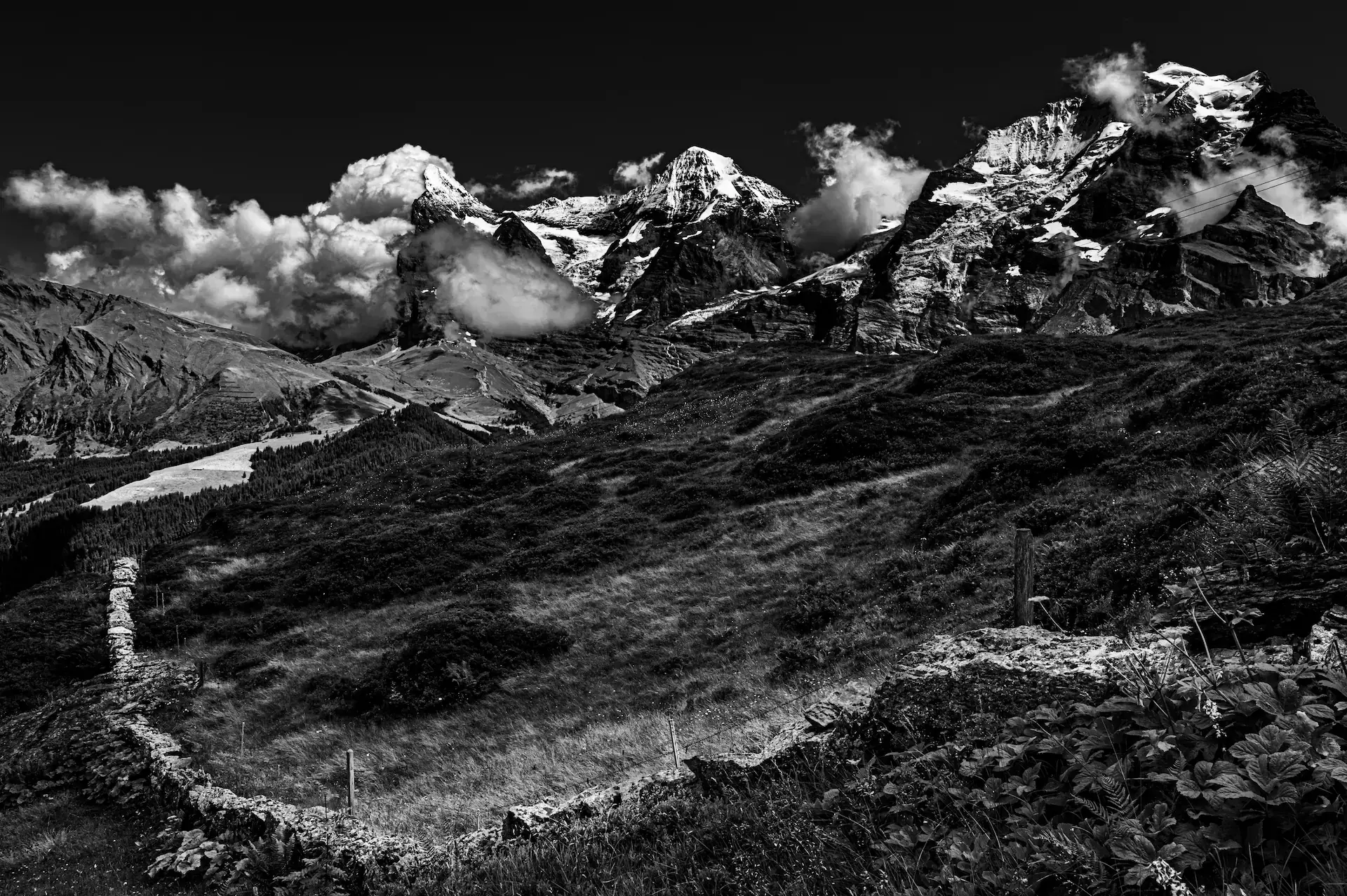 Black-and-white view of the Eiger, Mönch, and Jungfrau mountains above a stone wall near Wengen, Switzerland.