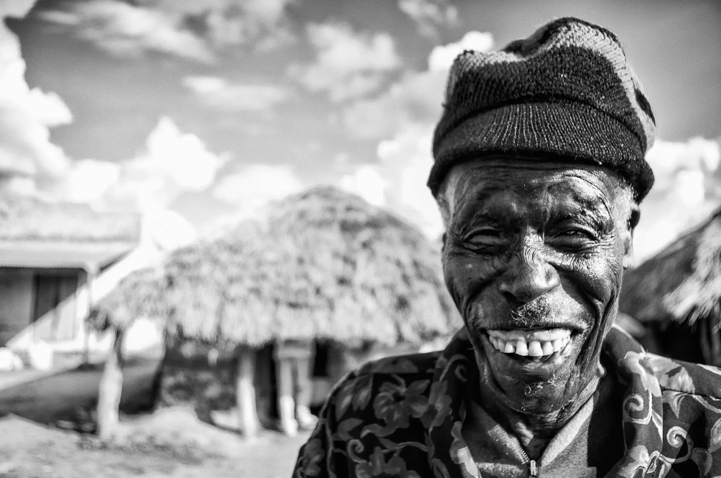 A Generous Smile Black-and-white close portrait of a smiling man wearing a knit cap and patterned shirt, with a thatched building and cloudy sky behind him in Uganda