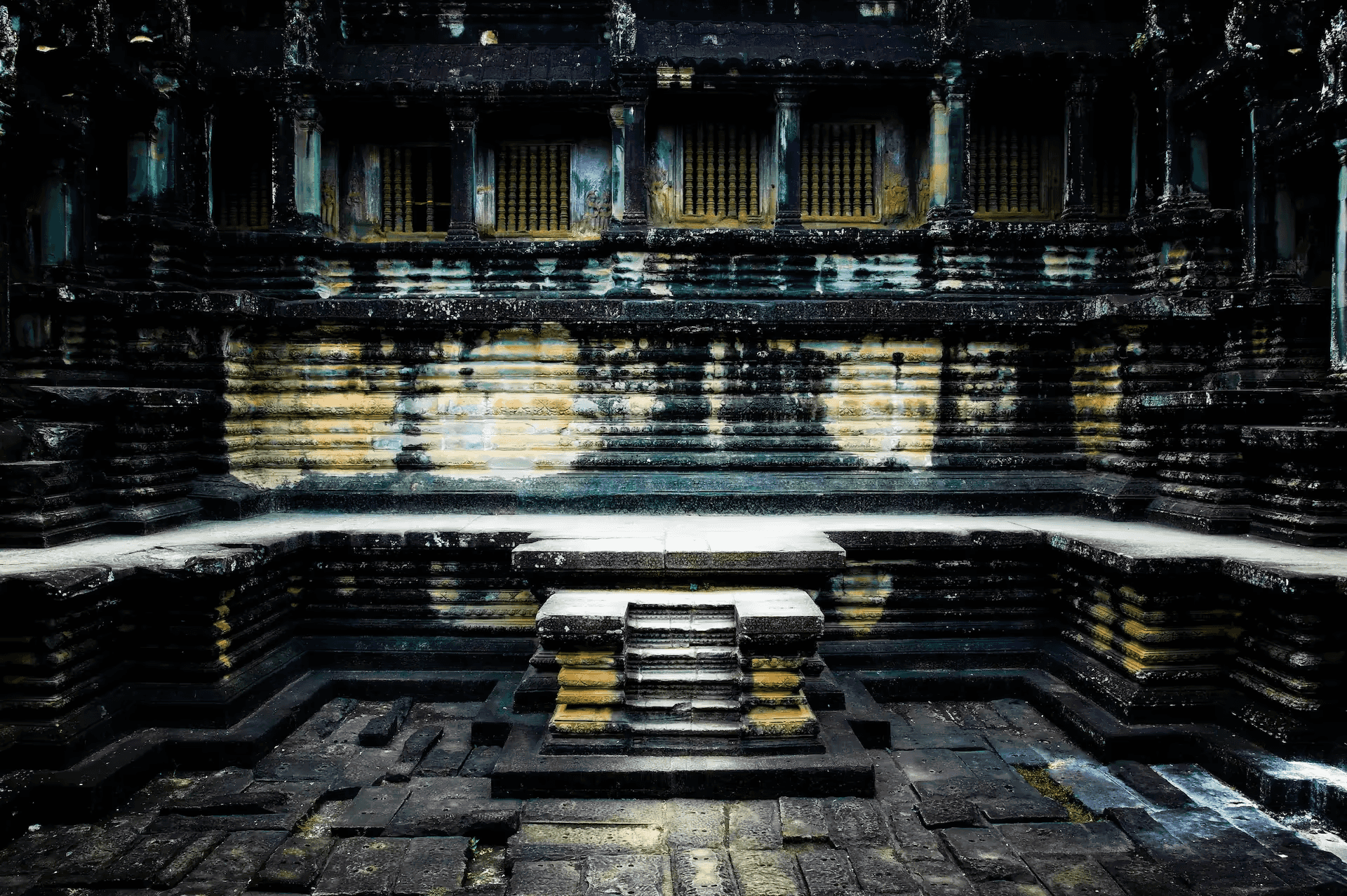 A detailed photograph showcasing the interior stone architecture of Angkor Wat temple in Cambodia. The image focuses on a section of the temple with layered stone walls, columns, and window openings with decorative latticework.