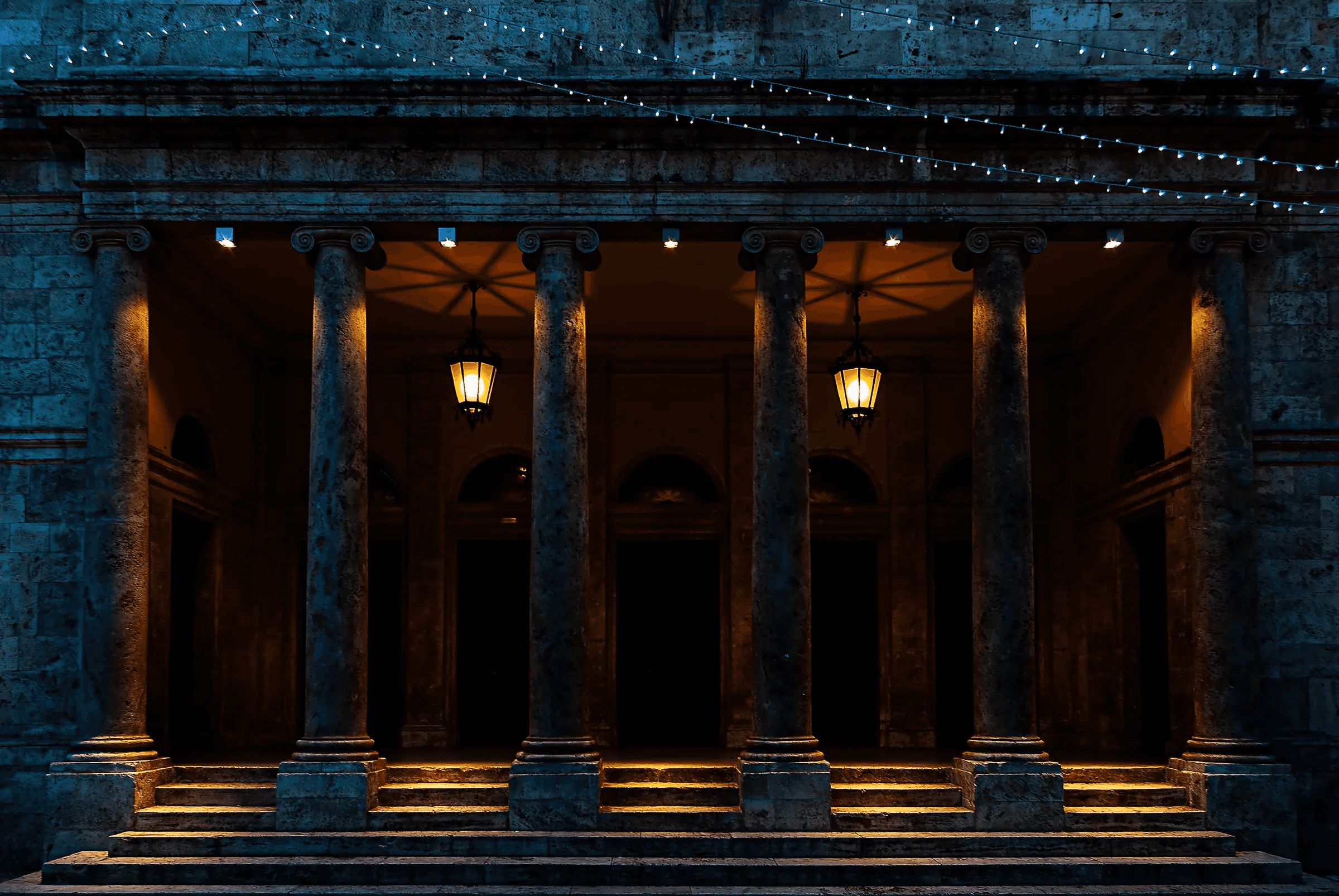 Color photograph taken at night showing the illuminated portico of a classical building in Ascoli Piceno, Italy, with columns, steps, hanging lanterns, and string lights.