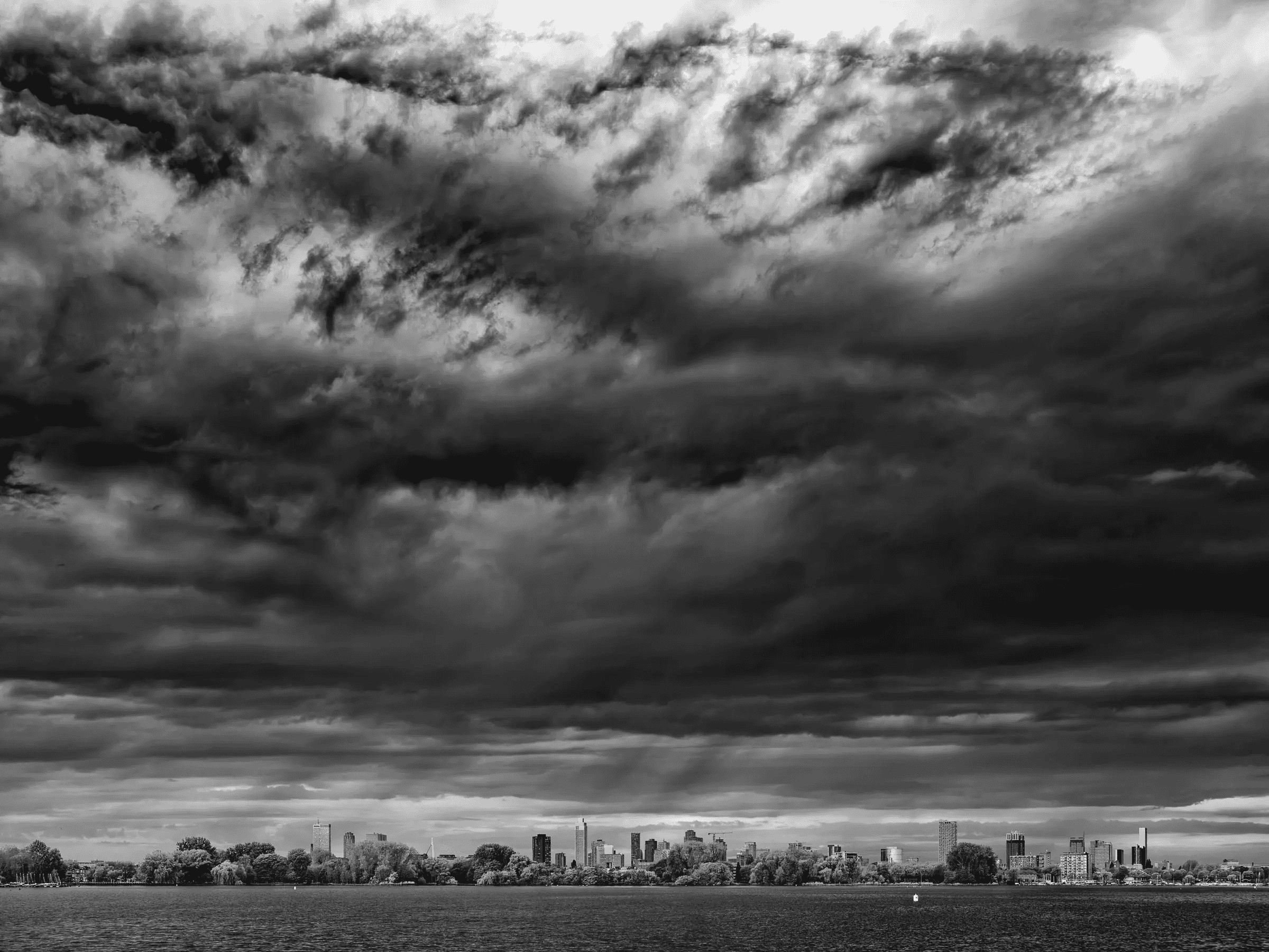 Brooding Skies Over the City Rotterdam Dramatic black and white photograph of a city skyline beneath heavy, textured storm clouds, seen from across a wide river or lake.