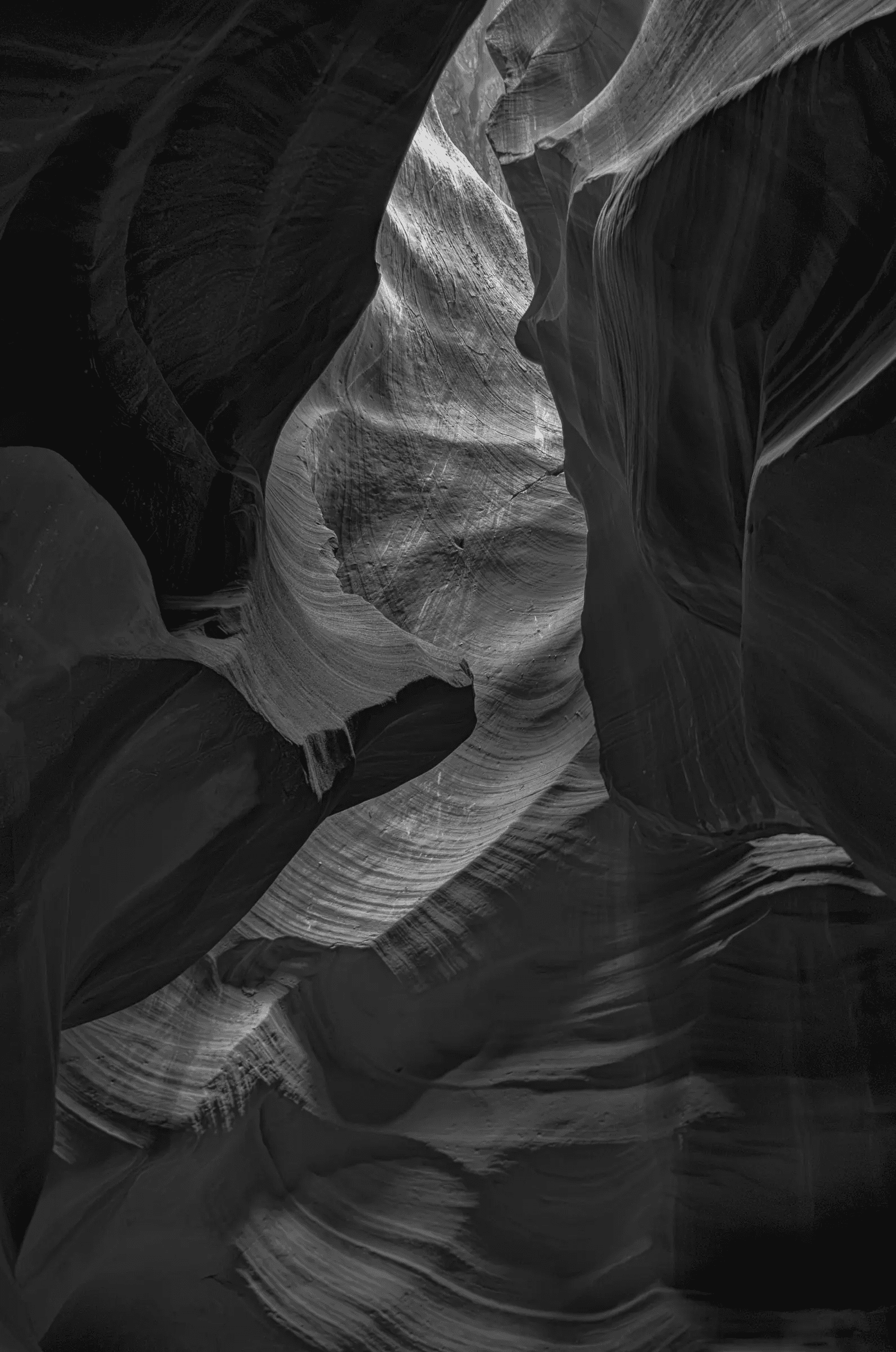 Interior view of a narrow slot canyon (Antelope Canyon) showing curved, layered sandstone walls and a shaft of light illuminating textured striations in black and white.