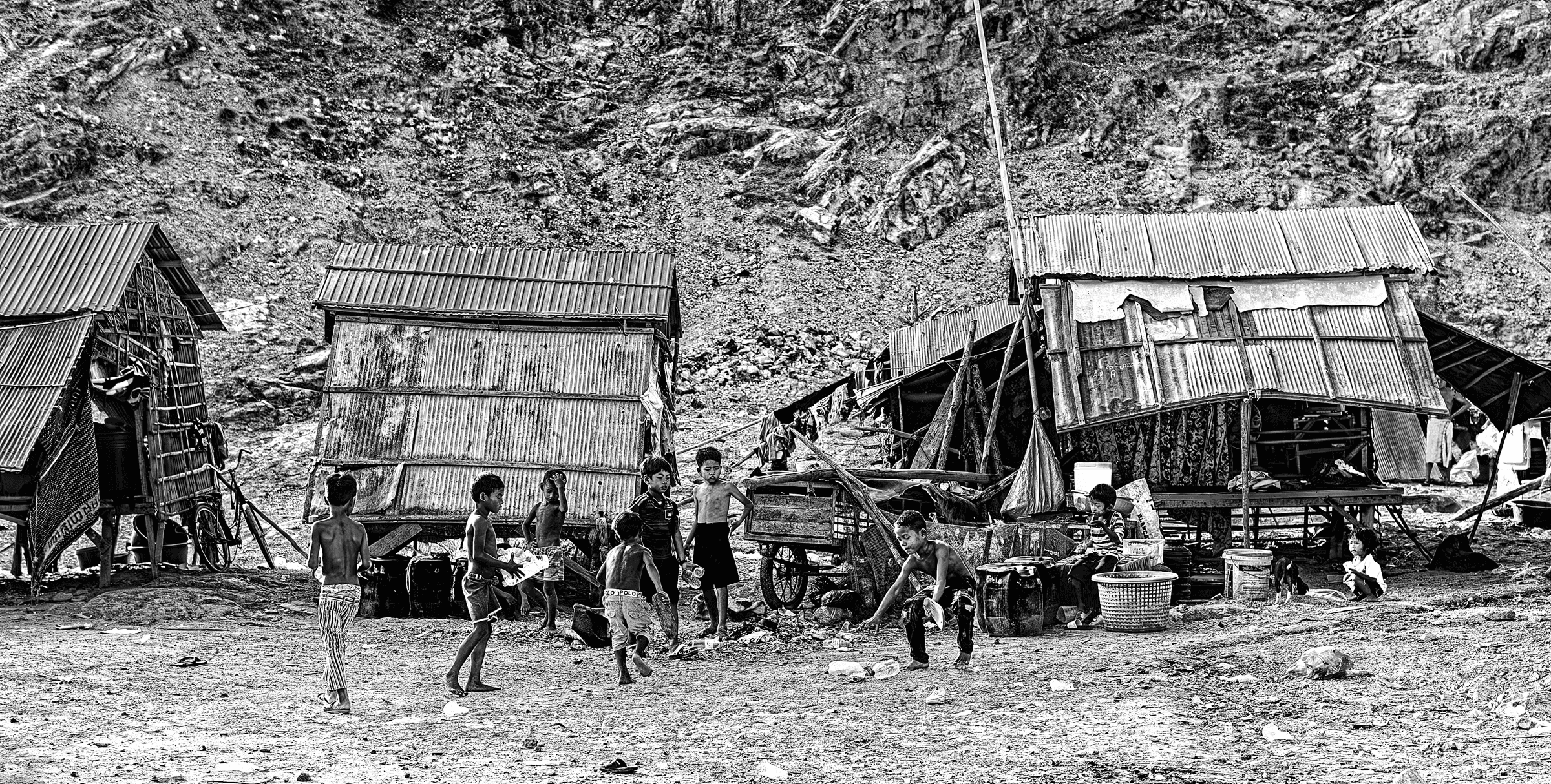 Black and white photograph of several children playing or standing outside simple corrugated metal structures at the base of a rocky hillside.