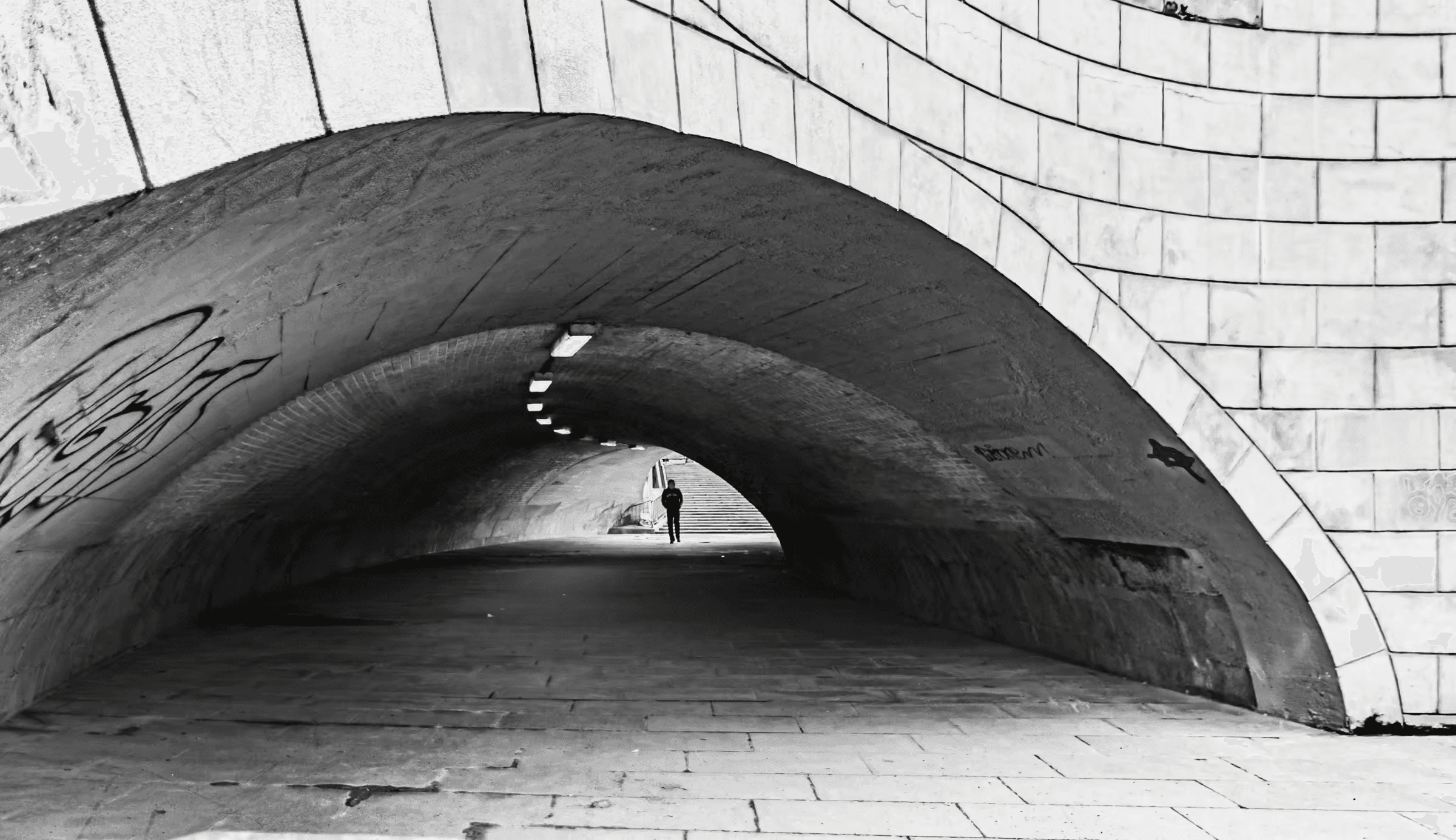 Black and white photograph of an arched city tunnel. The tunnel's brick or stone construction is visible in monochrome.