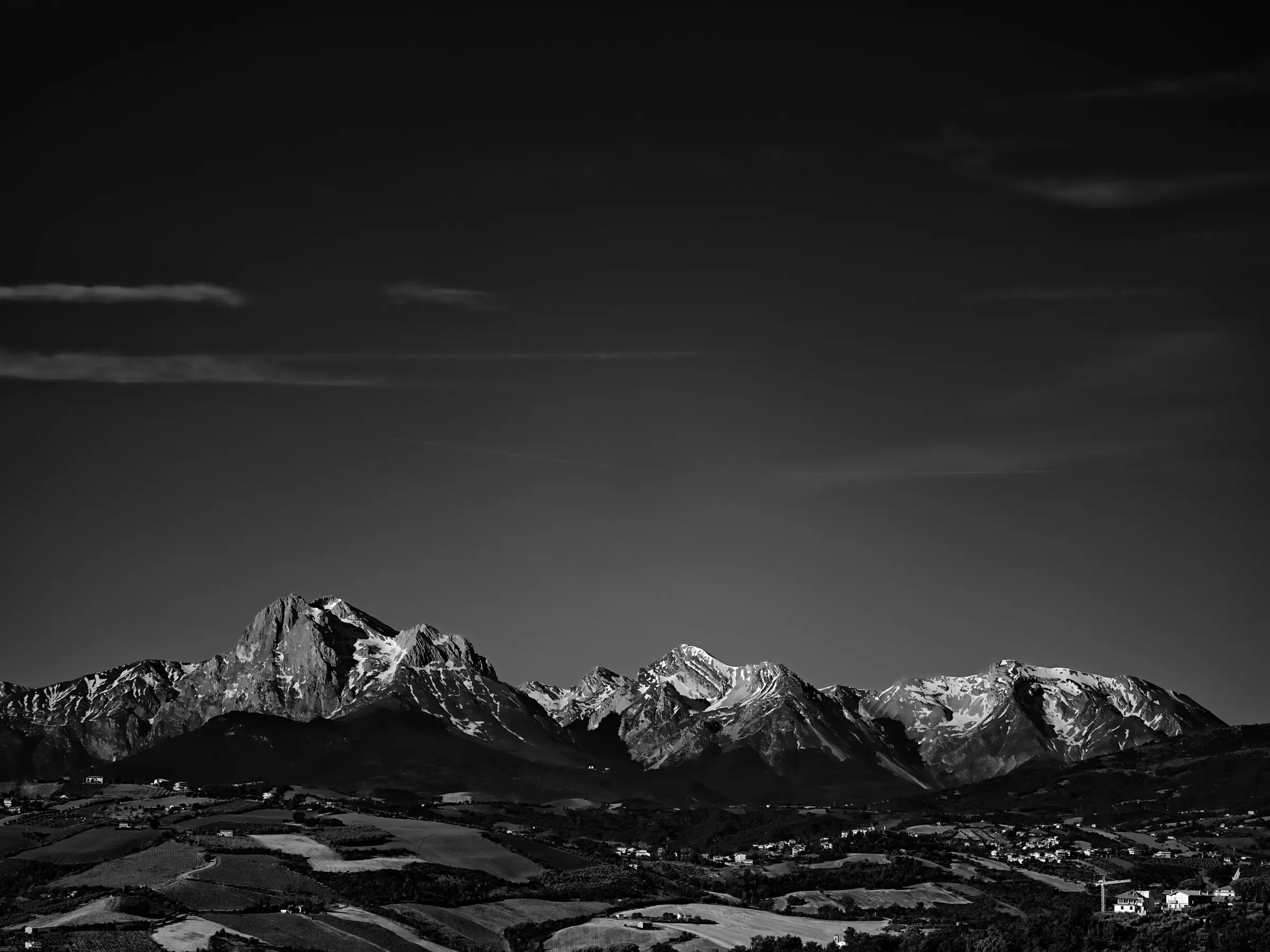 Dark Vault Black-and-white panoramic photograph of snow-covered mountains above a rural valley under a large dark sky with thin clouds.
