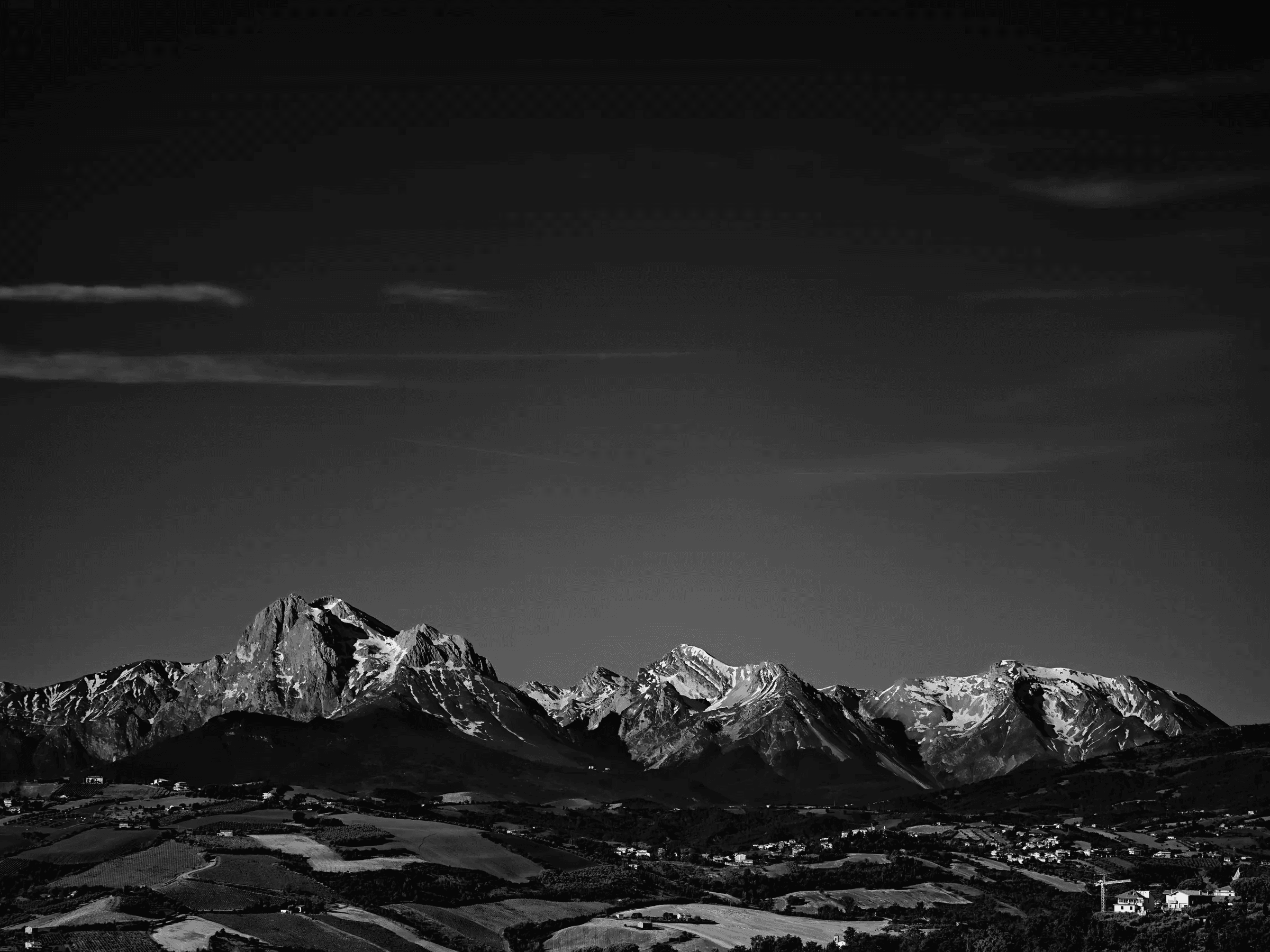 Dark Vault Black-and-white panoramic photograph of snow-covered mountains above a rural valley under a large dark sky with thin clouds.