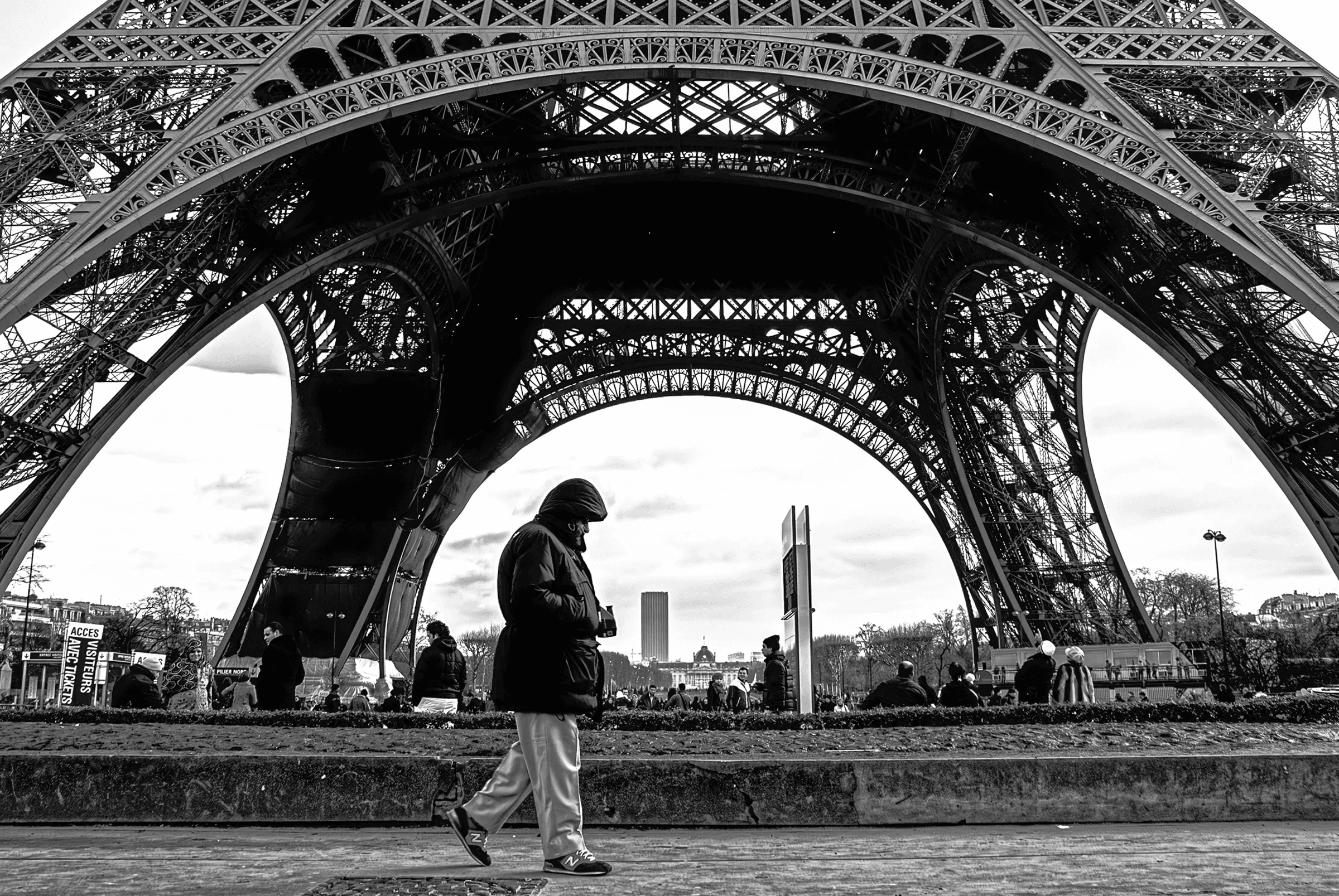 Black and white photograph of the Eiffel Tower in Paris, France, taken from a low angle.  A person is walking in the foreground, providing scale.
