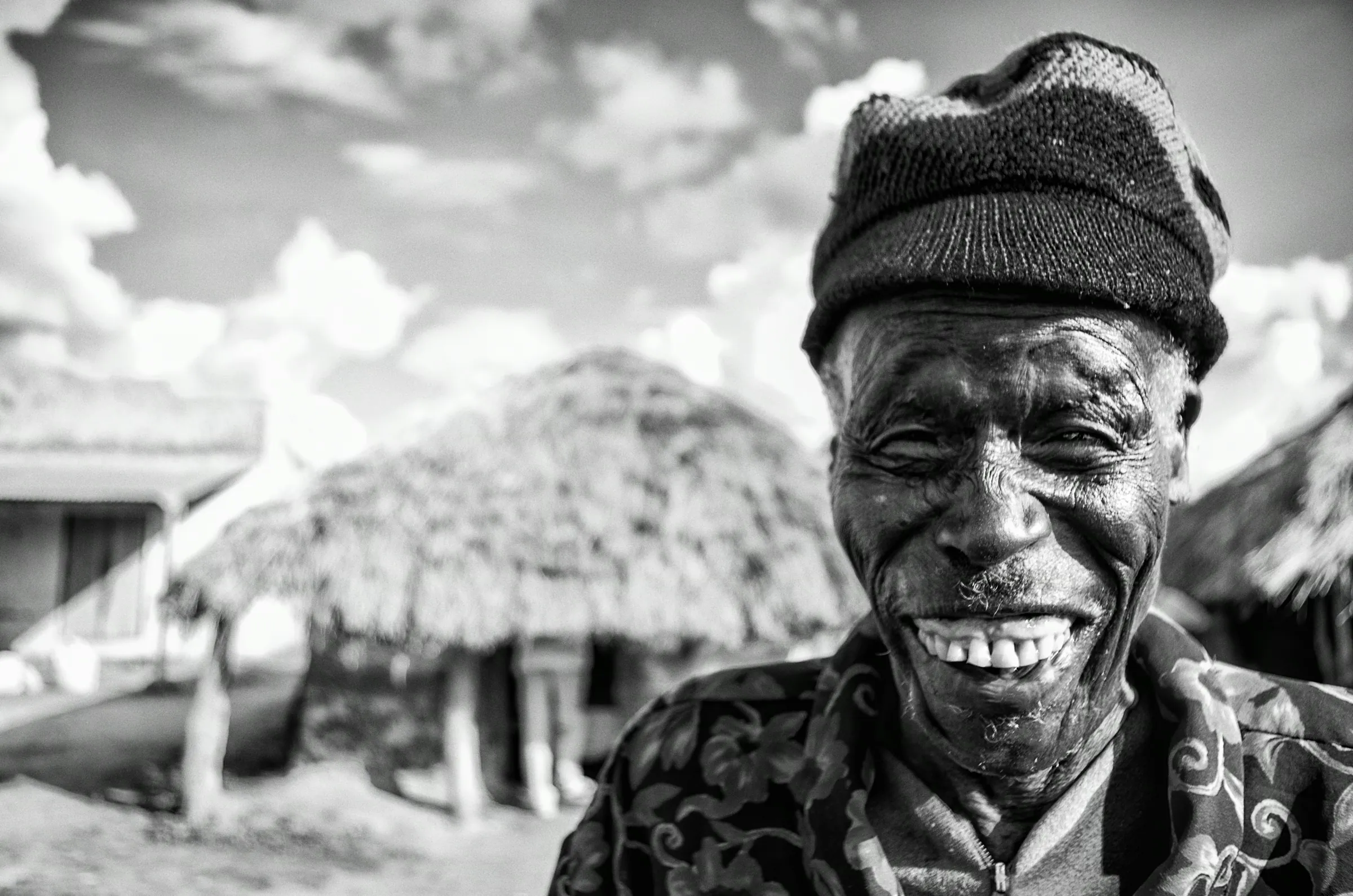 Black and white portrait of a smiling elderly man wearing a knit cap and patterned shirt, outdoors in Amuria, Uganda, with traditional thatched-roof huts blurred in the background..