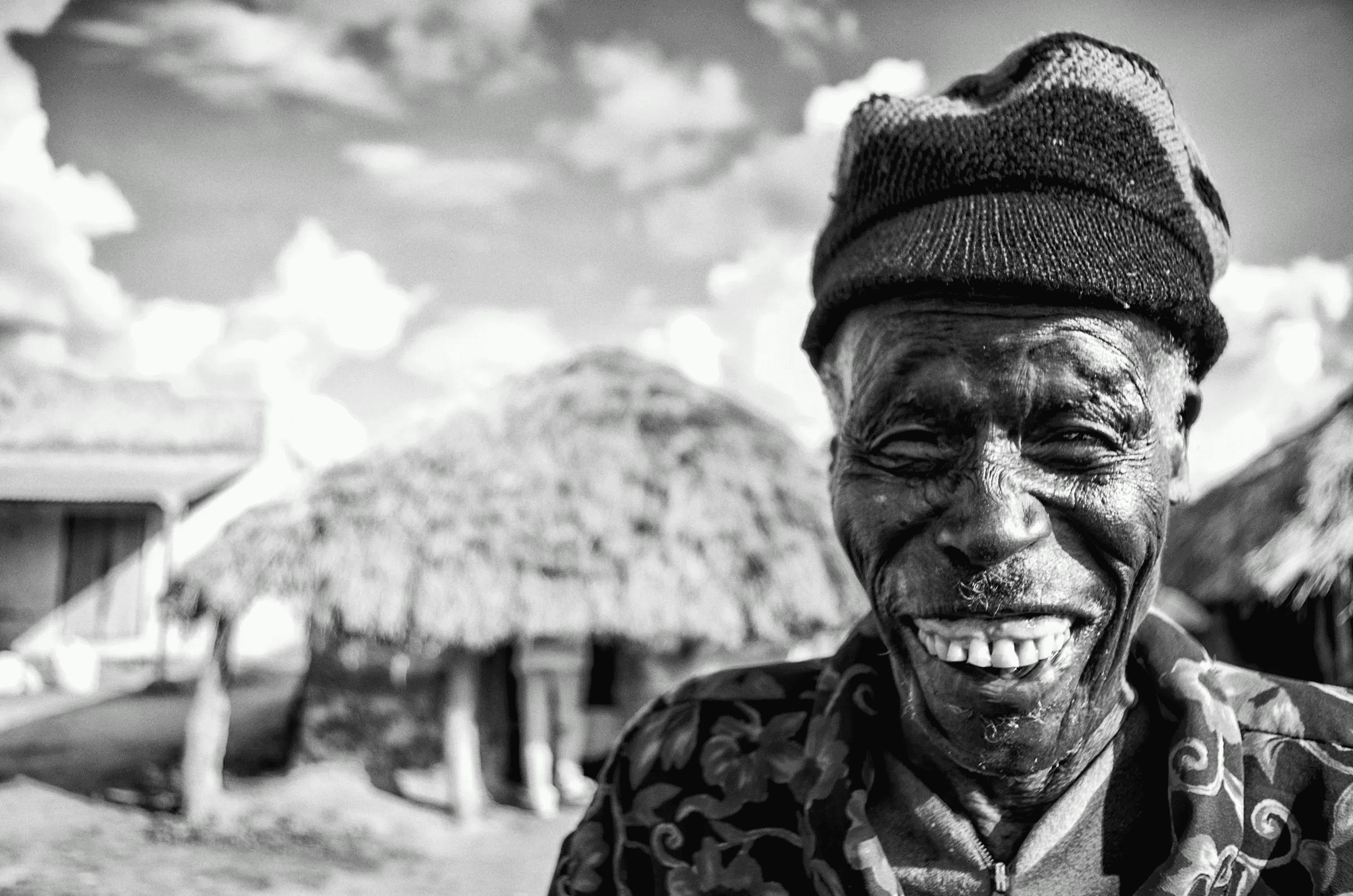 Black and white portrait of a smiling elderly man wearing a knit cap and patterned shirt, outdoors in Amuria, Uganda, with traditional thatched-roof huts blurred in the background..