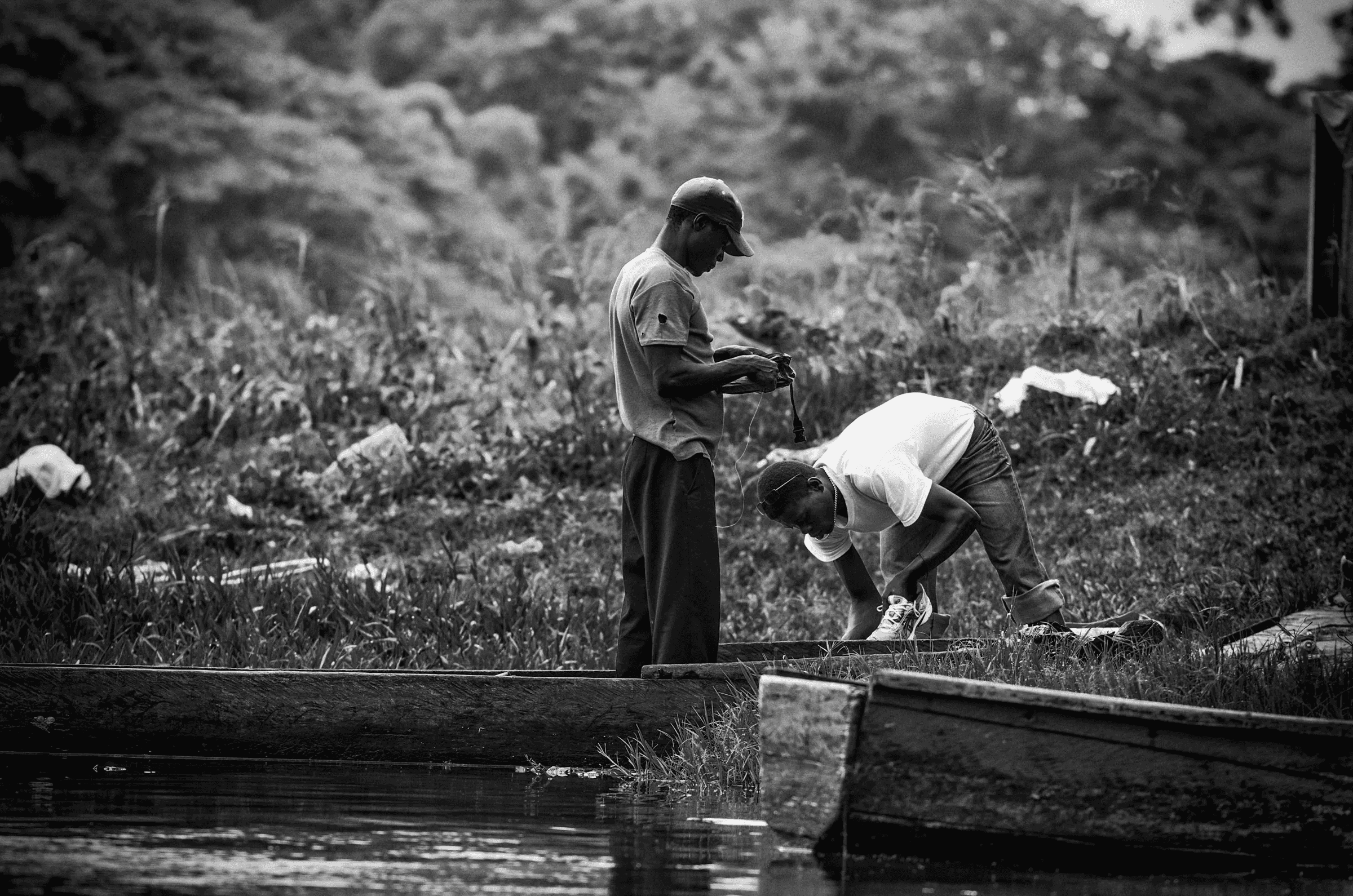 Black and white photograph showing two men working on or near simple boats/canoes on a riverbank, possibly preparing fishing gear.