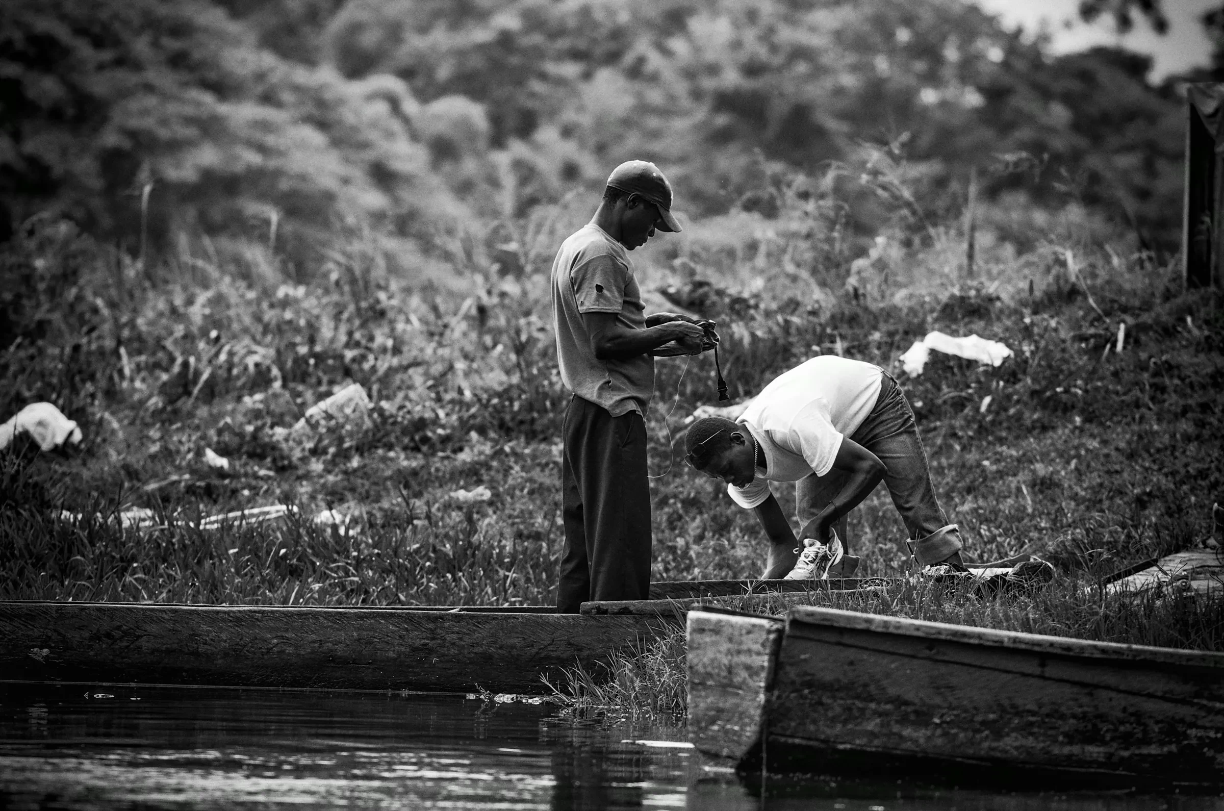 Two men working on or near simple boats/canoes on a riverbank, possibly preparing fishing gear.