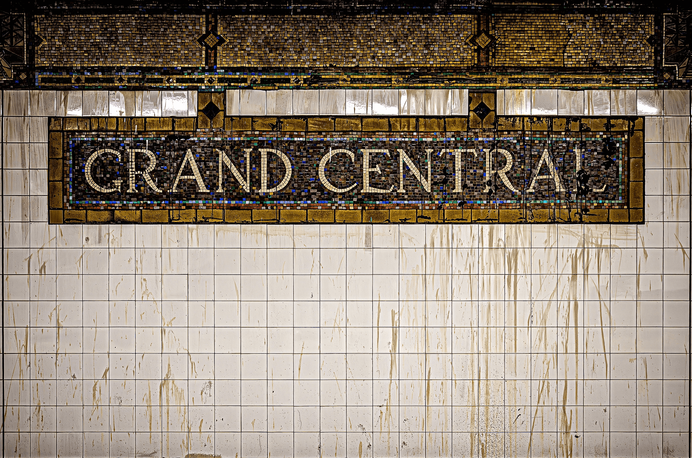 Color photograph close-up of the iconic mosaic tile sign reading 'GRAND CENTRAL' against aged white subway tiles in Grand Central Terminal, New York City.