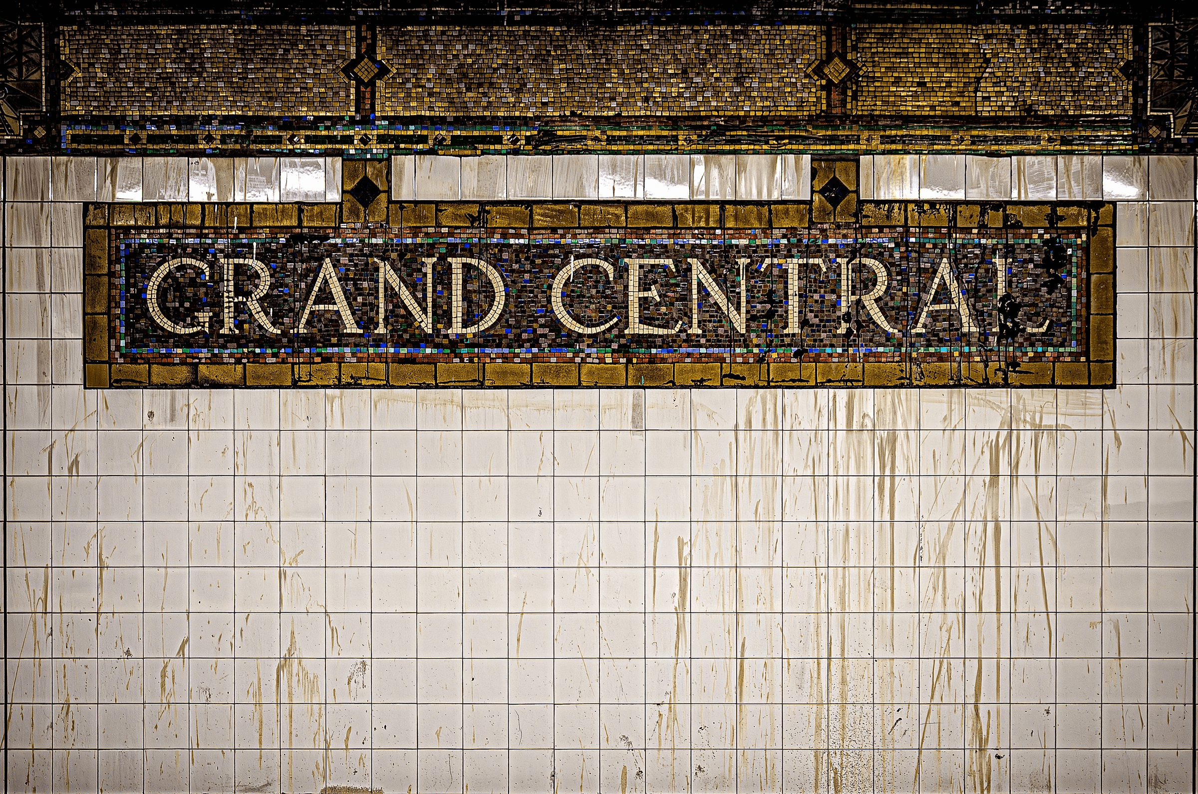 Color photograph close-up of the iconic mosaic tile sign reading 'GRAND CENTRAL' against aged white subway tiles in Grand Central Terminal, New York City.