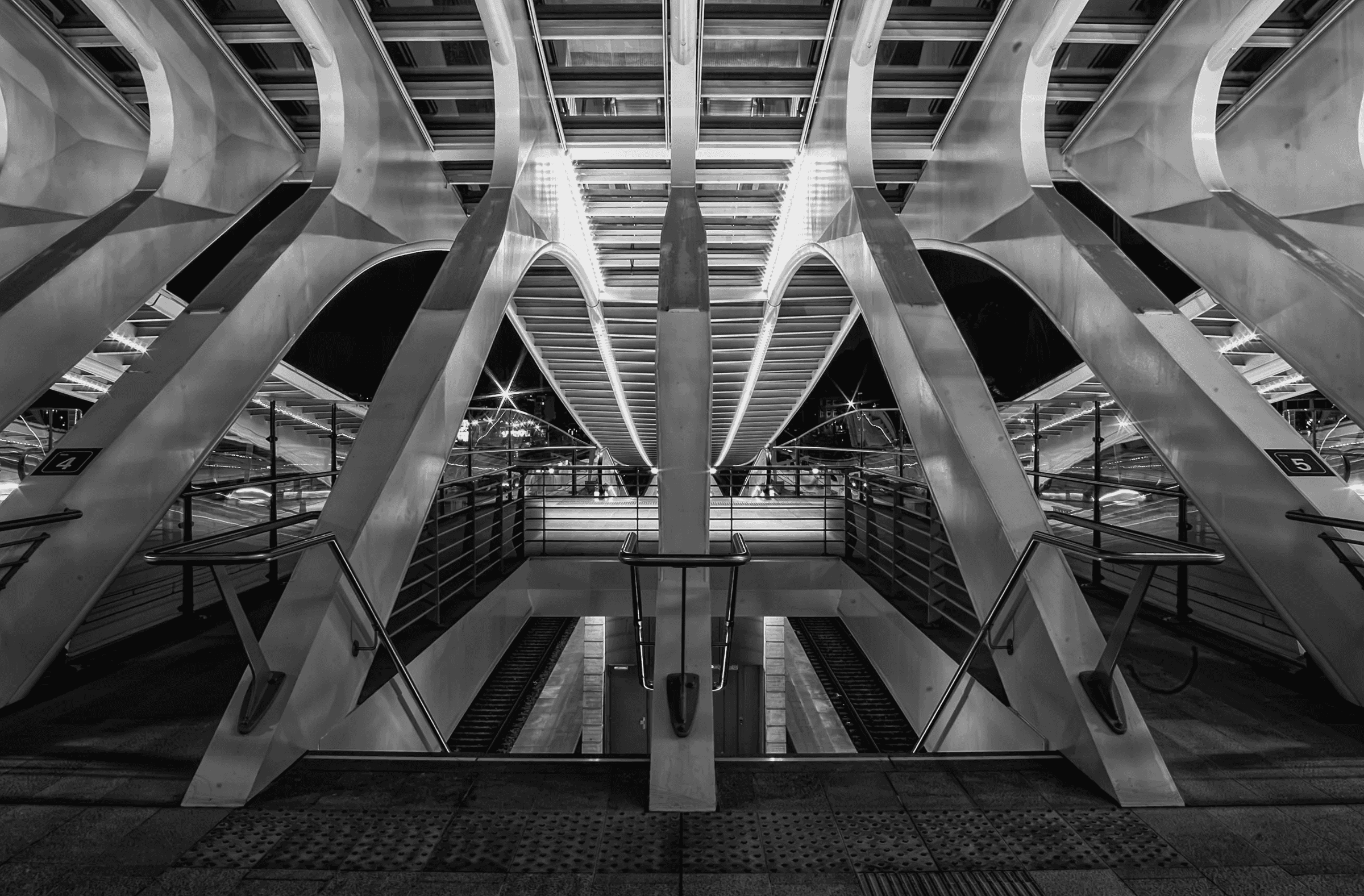 Symmetrical black and white photograph of the interior of Liège-Guillemins railway station, designed by Santiago Calatrava, showing the arched roof supports, platforms, and walkways.