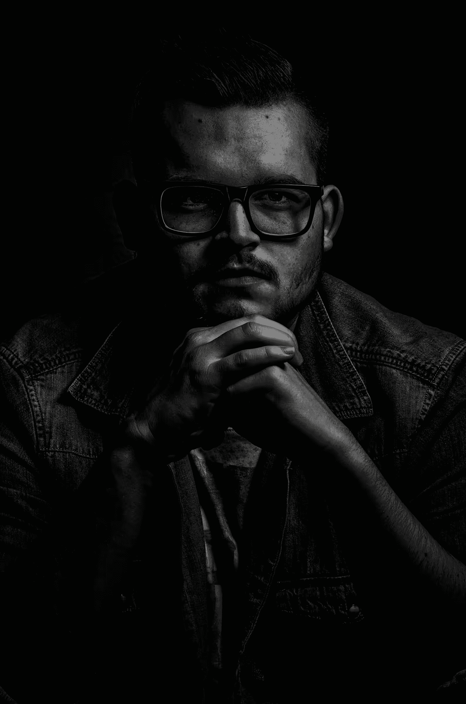 Contemplation in Shadow Low-key black and white portrait photograph of a man wearing glasses.