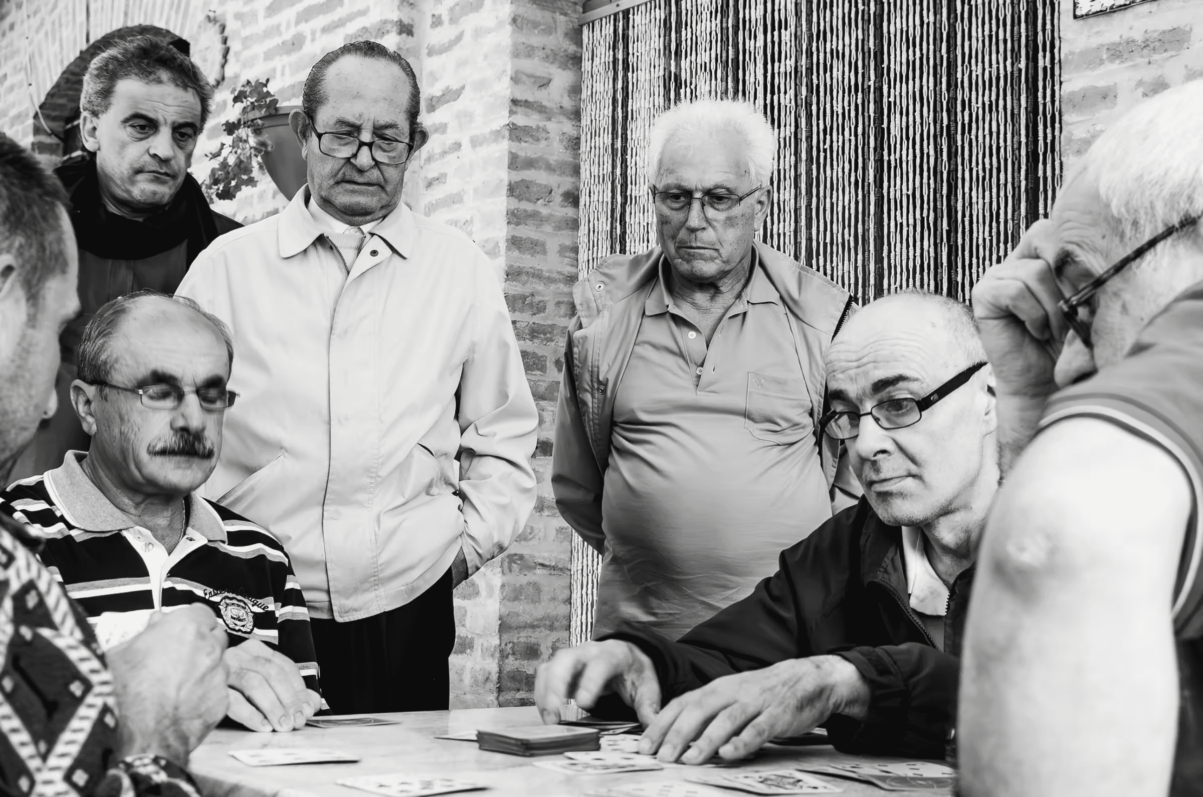 Black and white photograph of a group of older men playing cards. The men are intently focused on their cards and the game, their expressions serious and engaged.