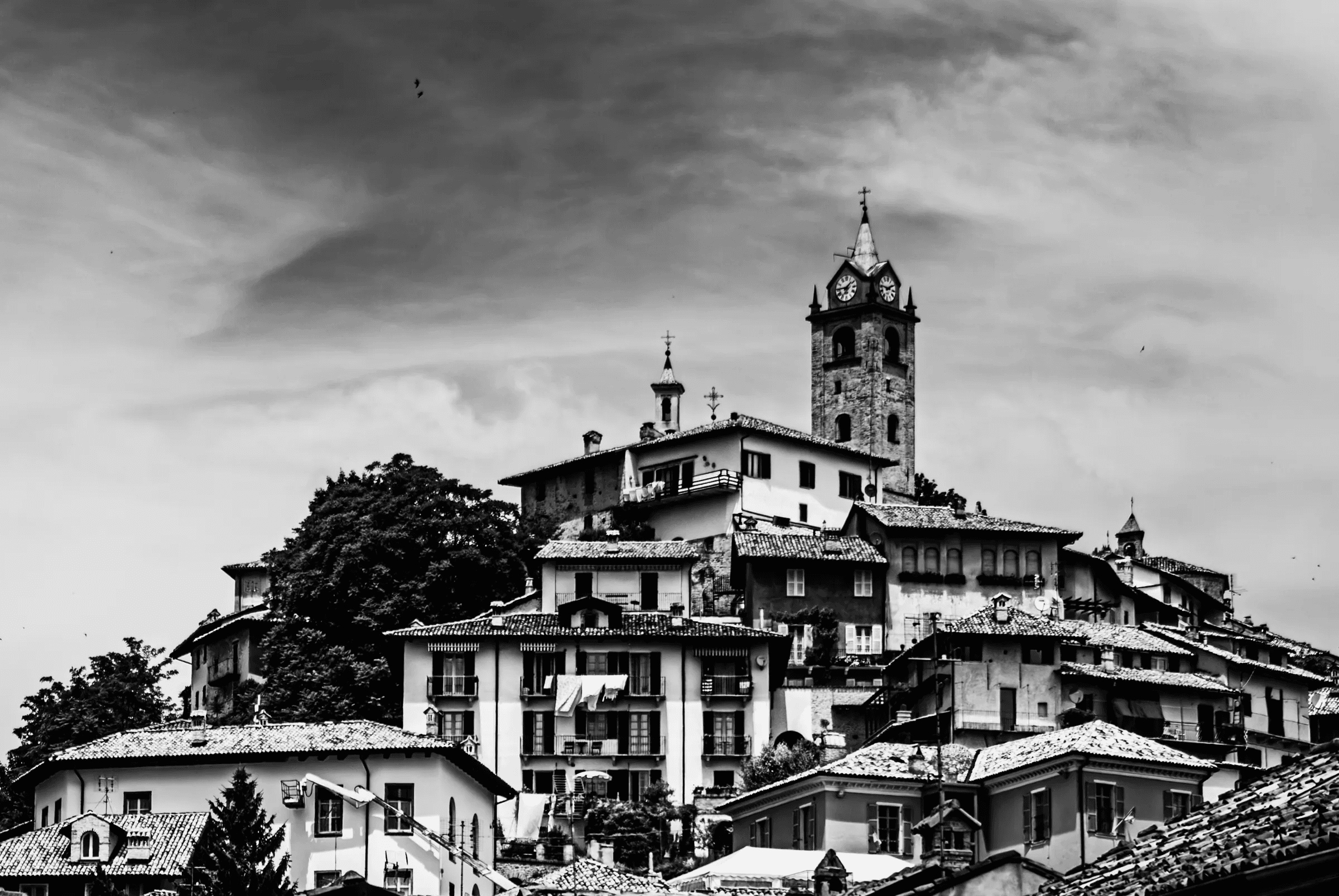 Black and white photograph looking up at the hilltop town of Monforte d'Alba, Piedmont, Italy, showing buildings clustered around a prominent clock tower under a dramatic sky.