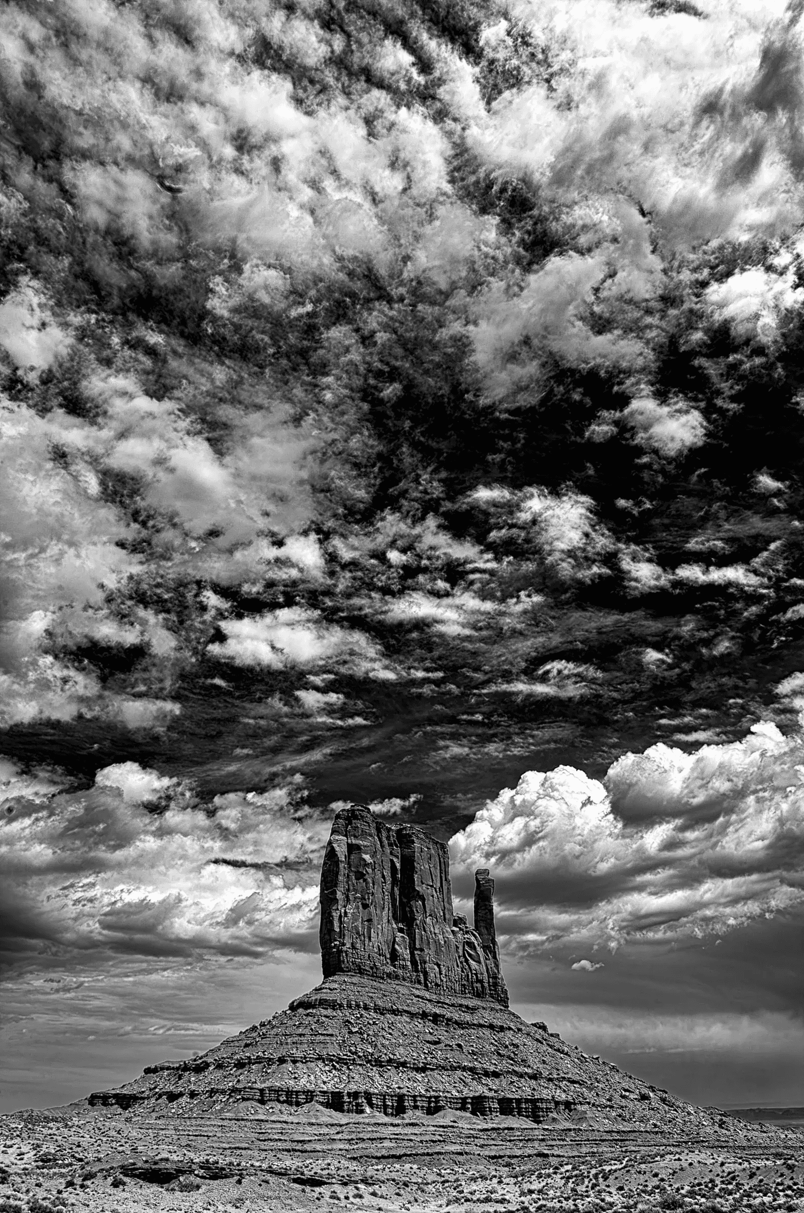 Black and white photograph of a solitary sandstone butte in Monument Valley beneath a dramatic, textured sky filled with layered clouds.