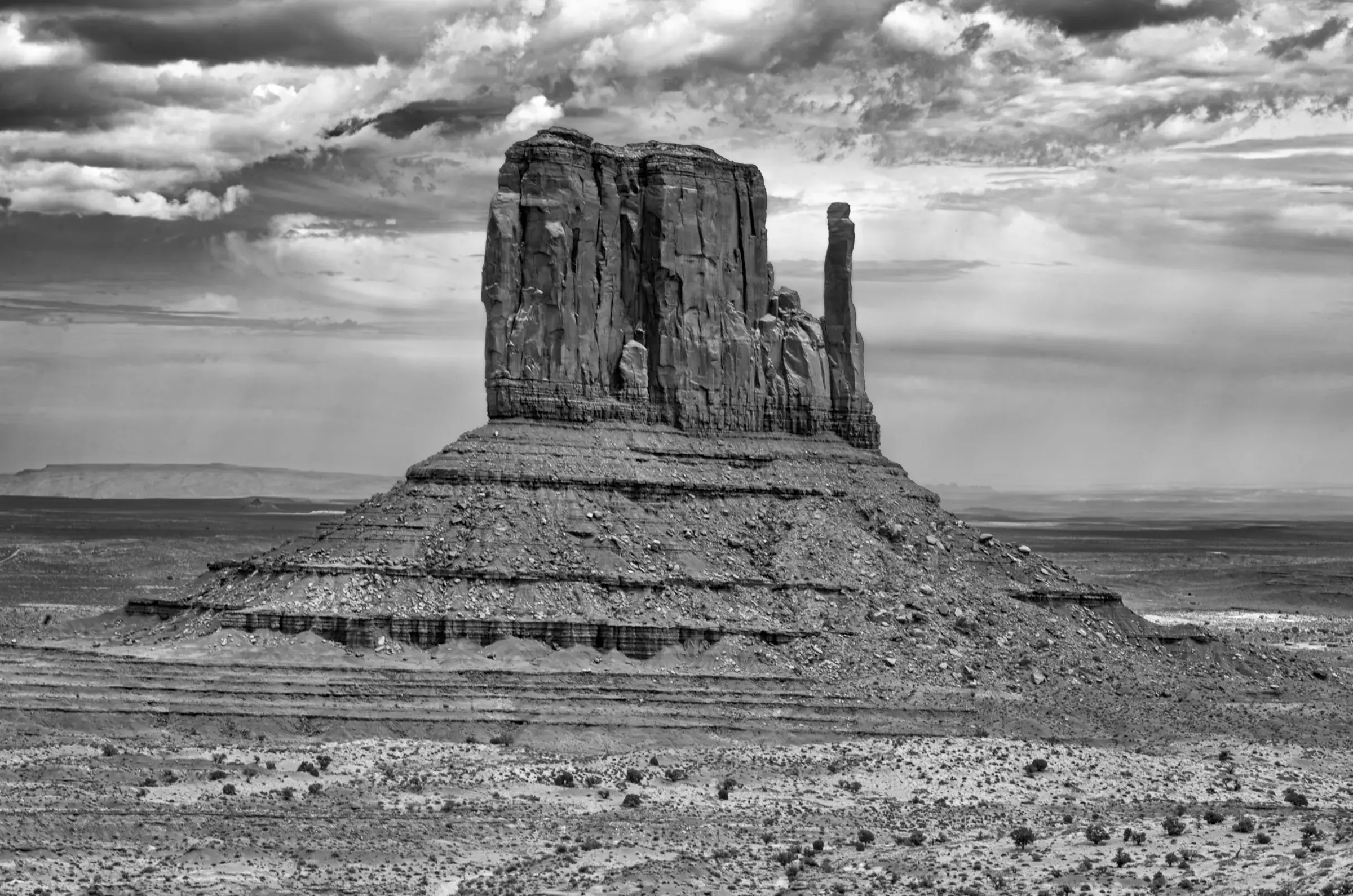 Monolith, Monument Valley A massive sandstone butte stands alone in a wide desert plain under dramatic, cloud-filled skies. The image is in black and white.