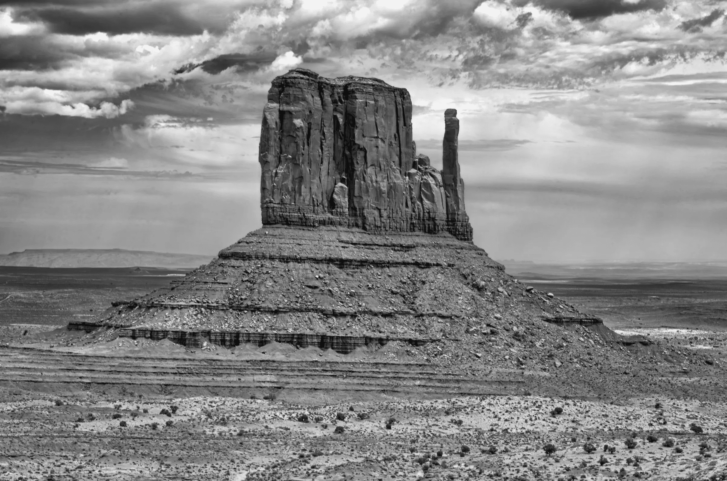 Monolith, Monument Valley A massive sandstone butte stands alone in a wide desert plain under dramatic, cloud-filled skies. The image is in black and white.
