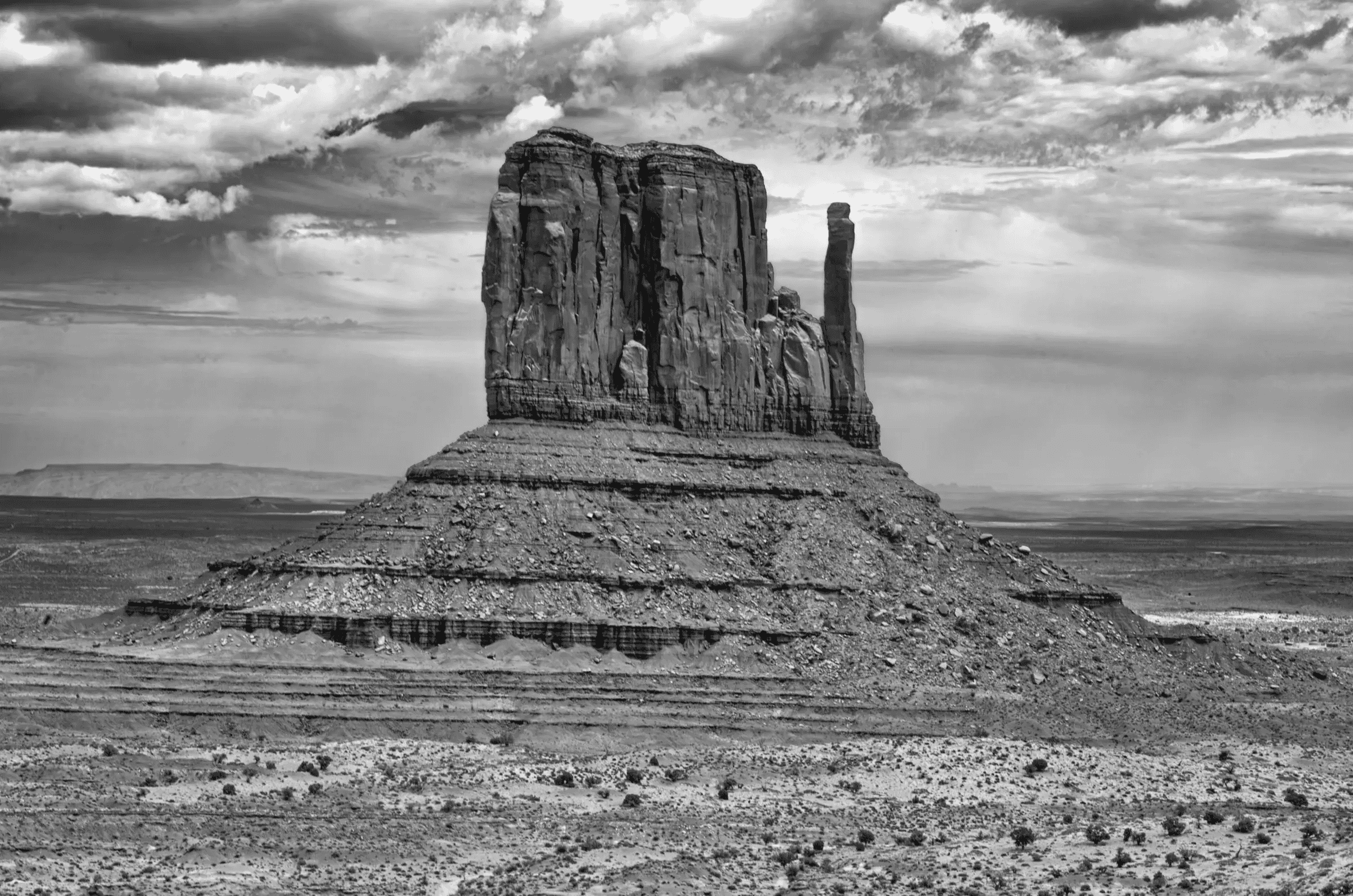 Monolith, Monument Valley A massive sandstone butte stands alone in a wide desert plain under dramatic, cloud-filled skies. The image is in black and white.