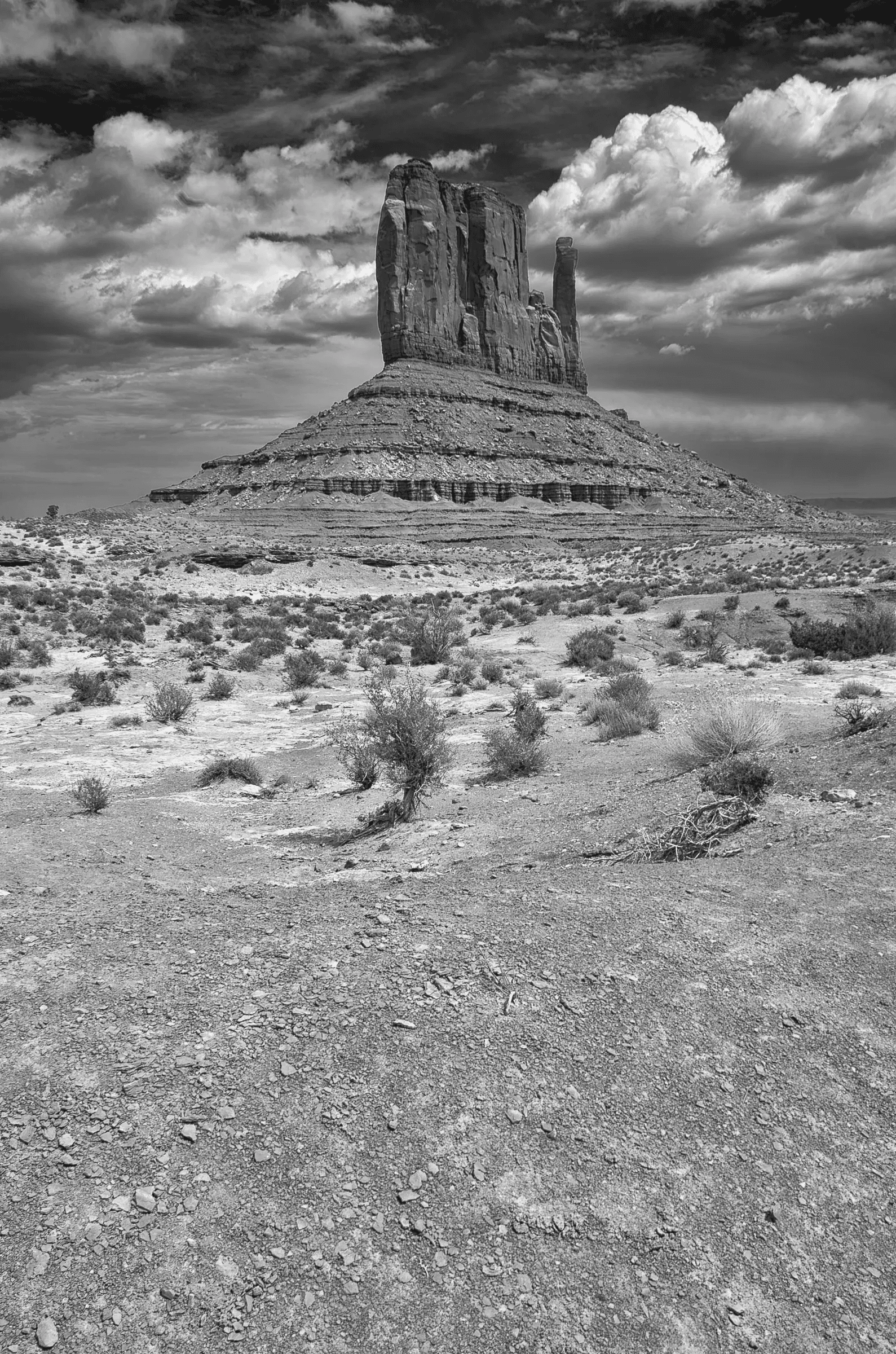 Monolith, Monument Valley A black and white photograph of a large sandstone butte in Monument Valley, with dramatic clouds overhead and sparse desert vegetation in the foreground.