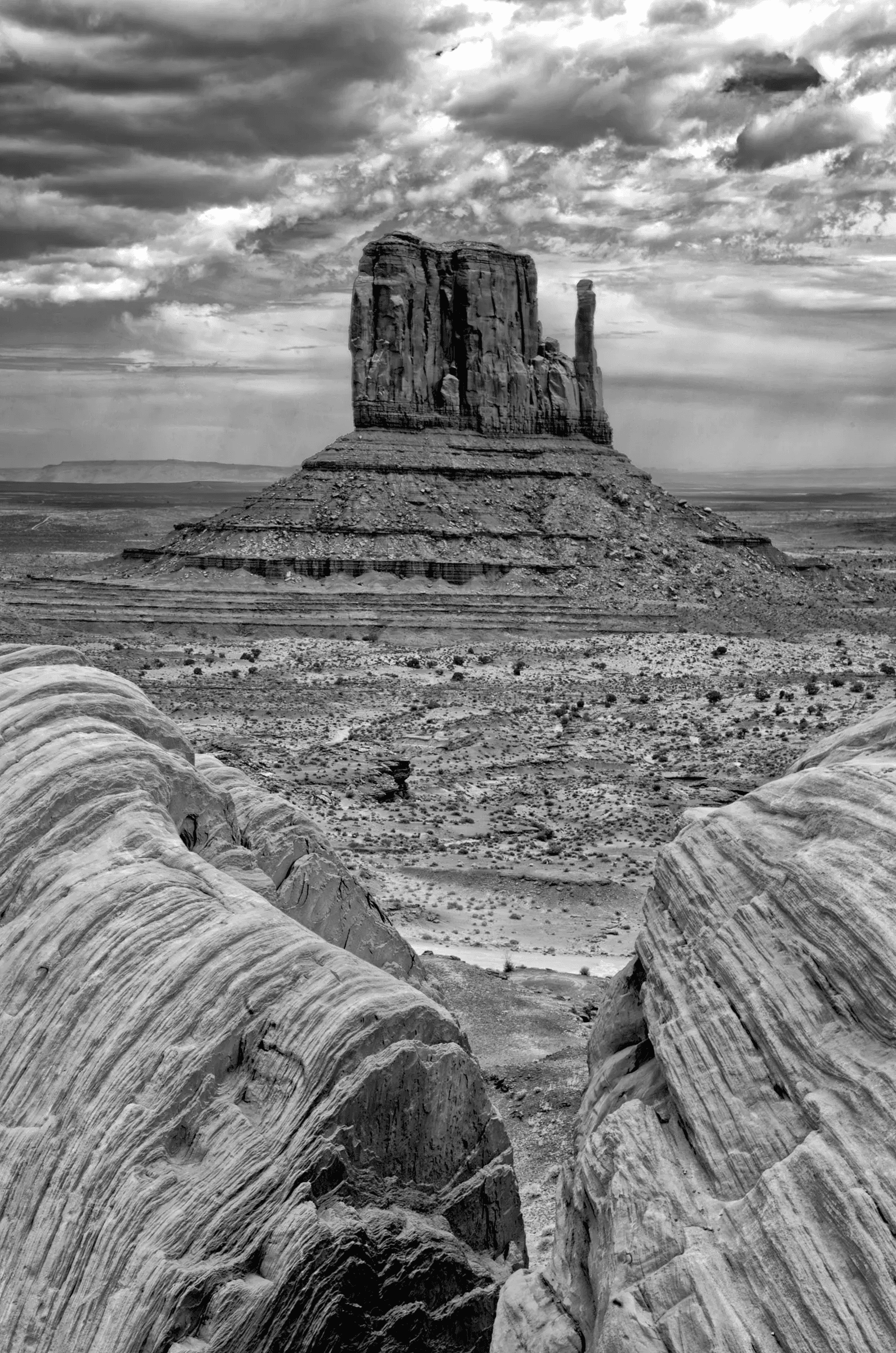 Monolith, Under Gathering Sky Black and white photograph of a large sandstone butte in a desert landscape, framed by textured foreground rocks and dramatic, cloud-filled sky.