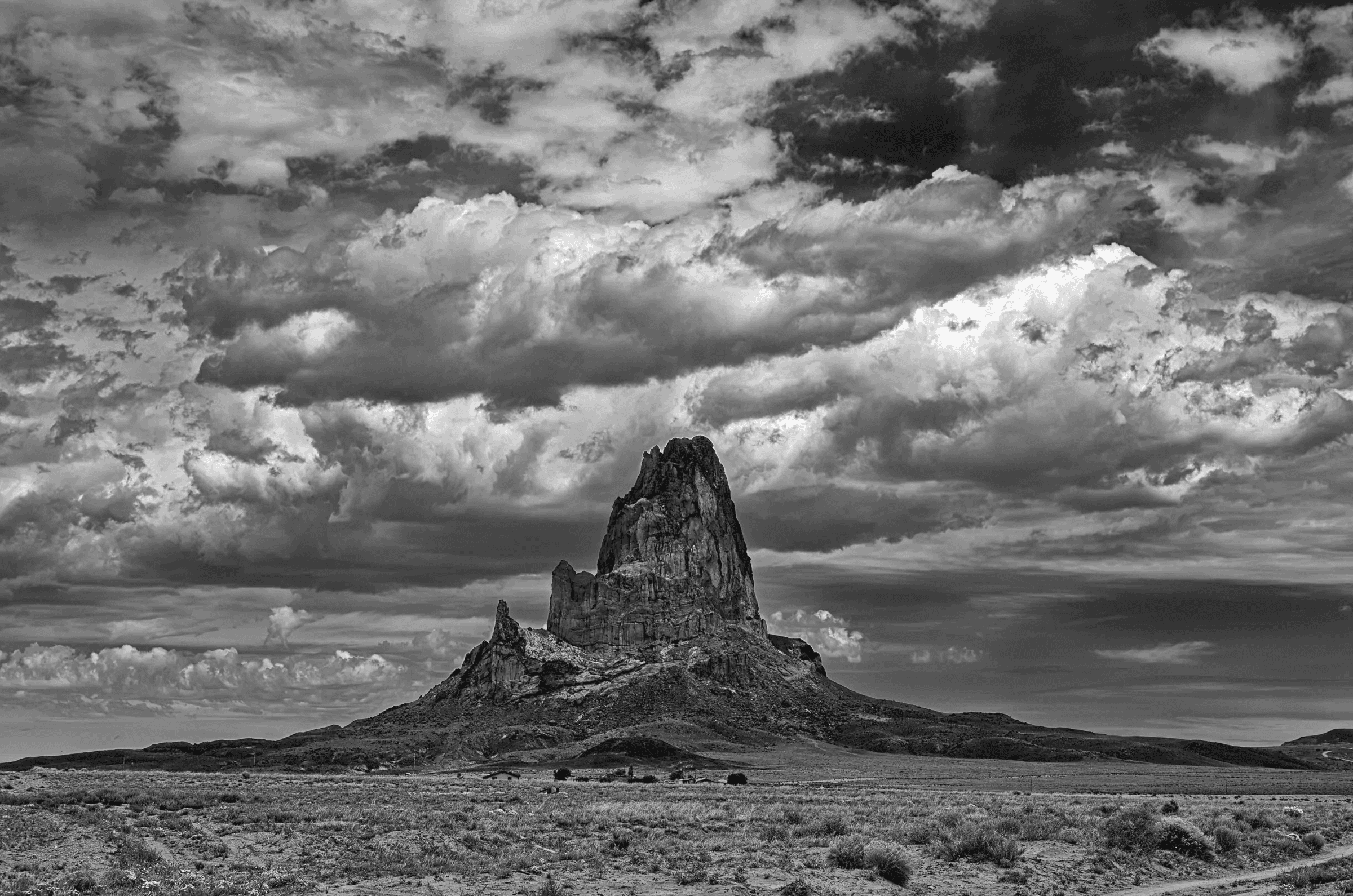Monolith Under Gathering Sky A dramatic black and white photograph of a towering rock formation rising from arid plains beneath a vast, textured sky filled with heavy, swirling clouds.