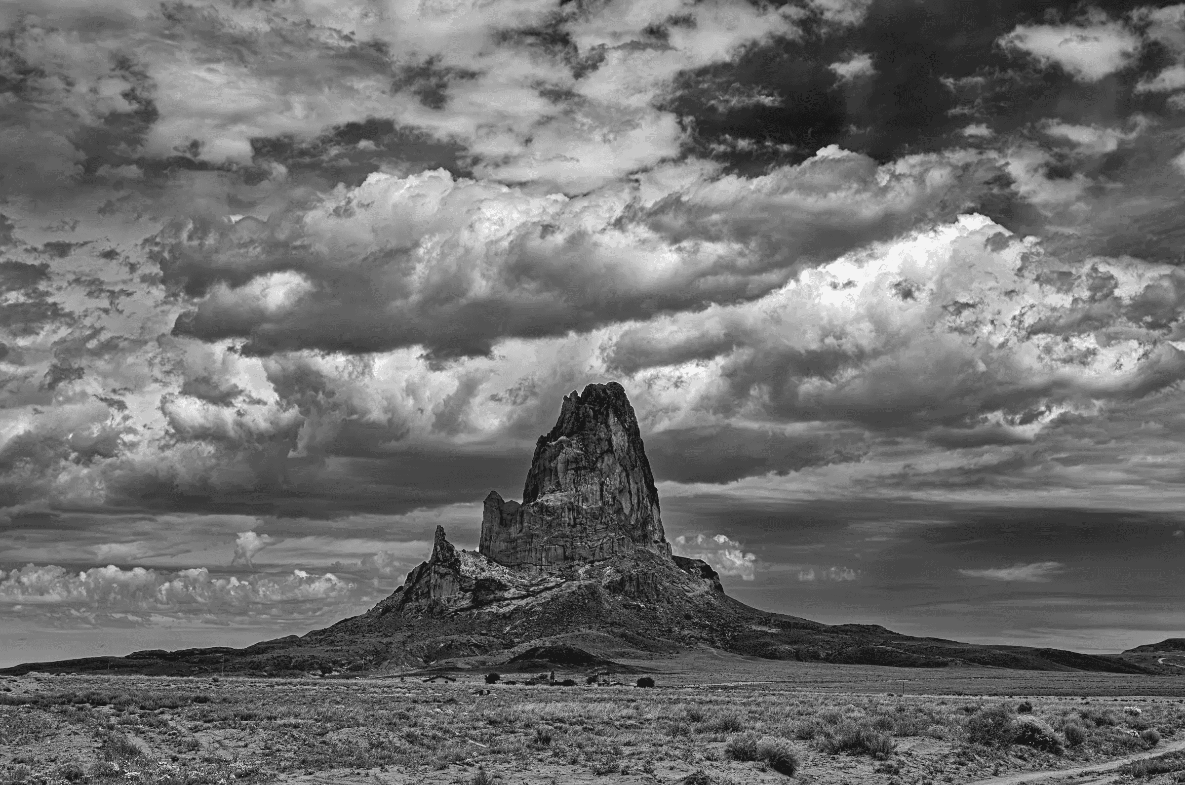 Monolith Under Gathering Sky A dramatic black and white photograph of a towering rock formation rising from arid plains beneath a vast, textured sky filled with heavy, swirling clouds.