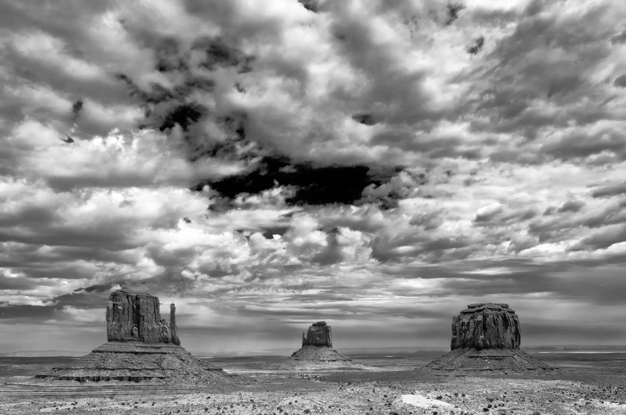 Monumental Weather Three large rock formations rise from a flat desert plain beneath a dramatic, cloud-filled sky in black and white.