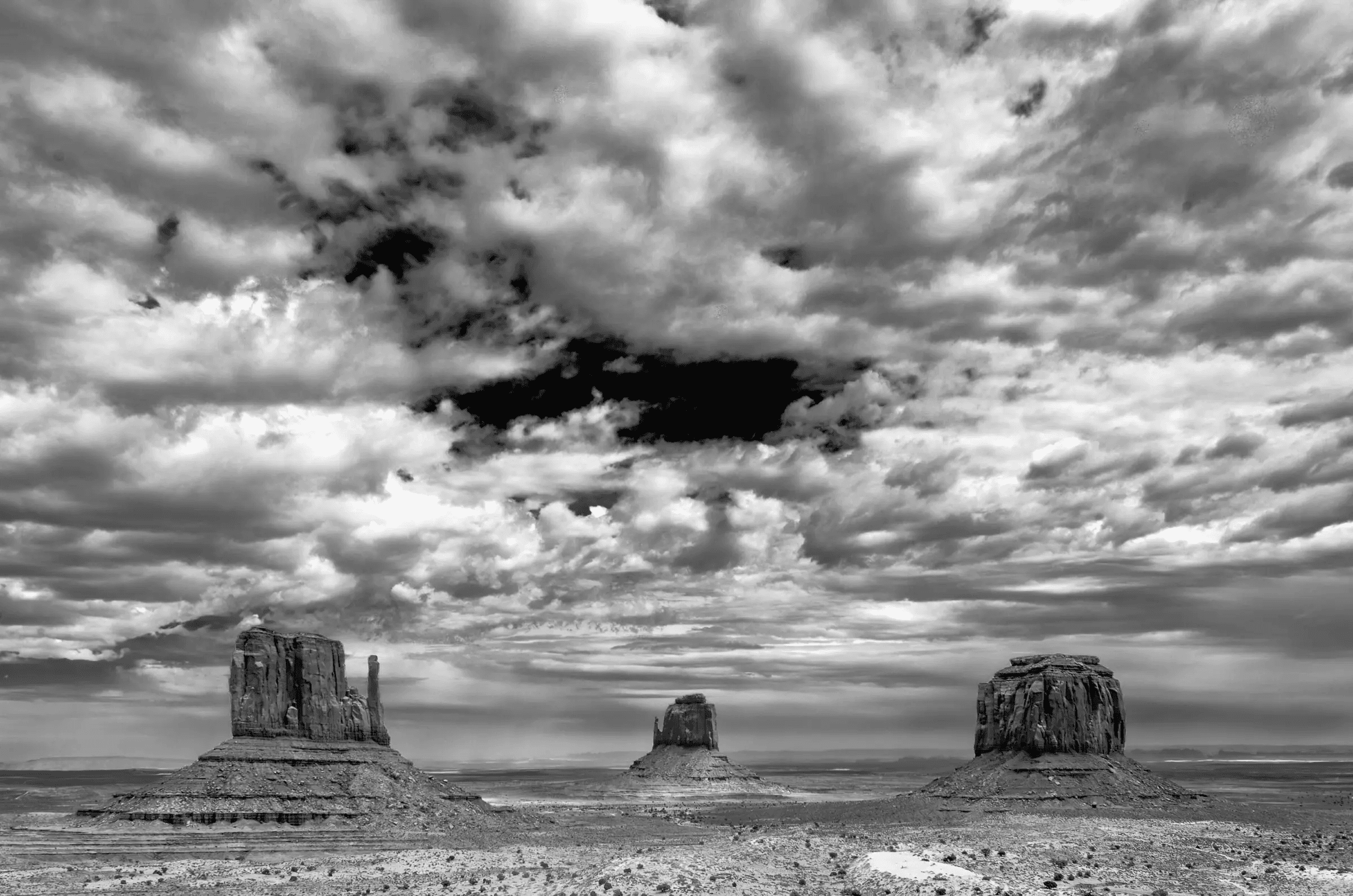 Monumental Weather Three large rock formations rise from a flat desert plain beneath a dramatic, cloud-filled sky in black and white.