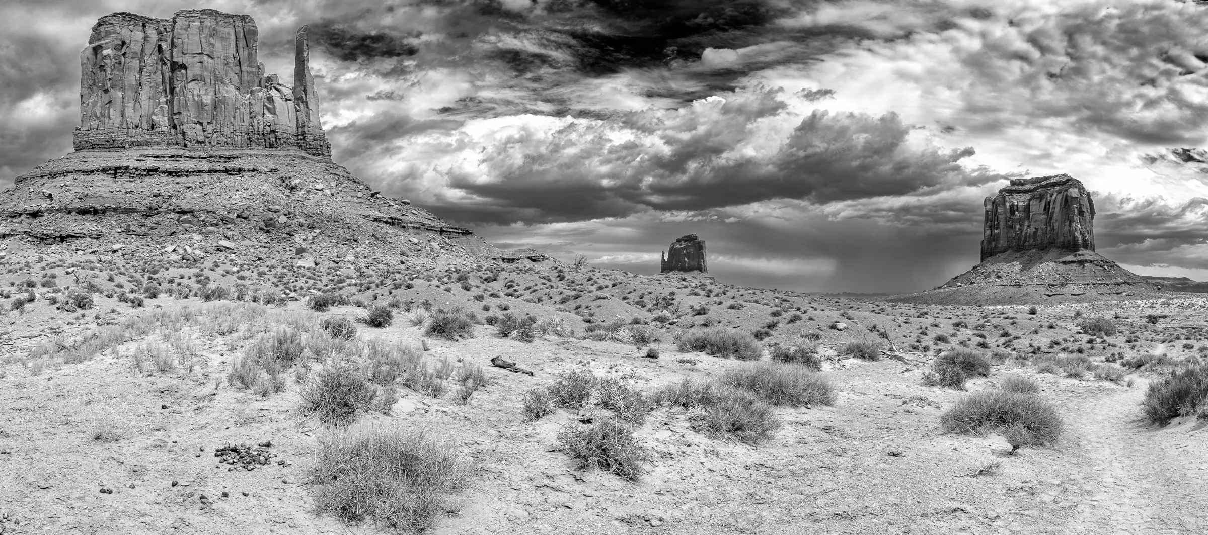 Black and white panorama of Monument Valley with three large buttes rising from arid desert, beneath dramatic, textured clouds.