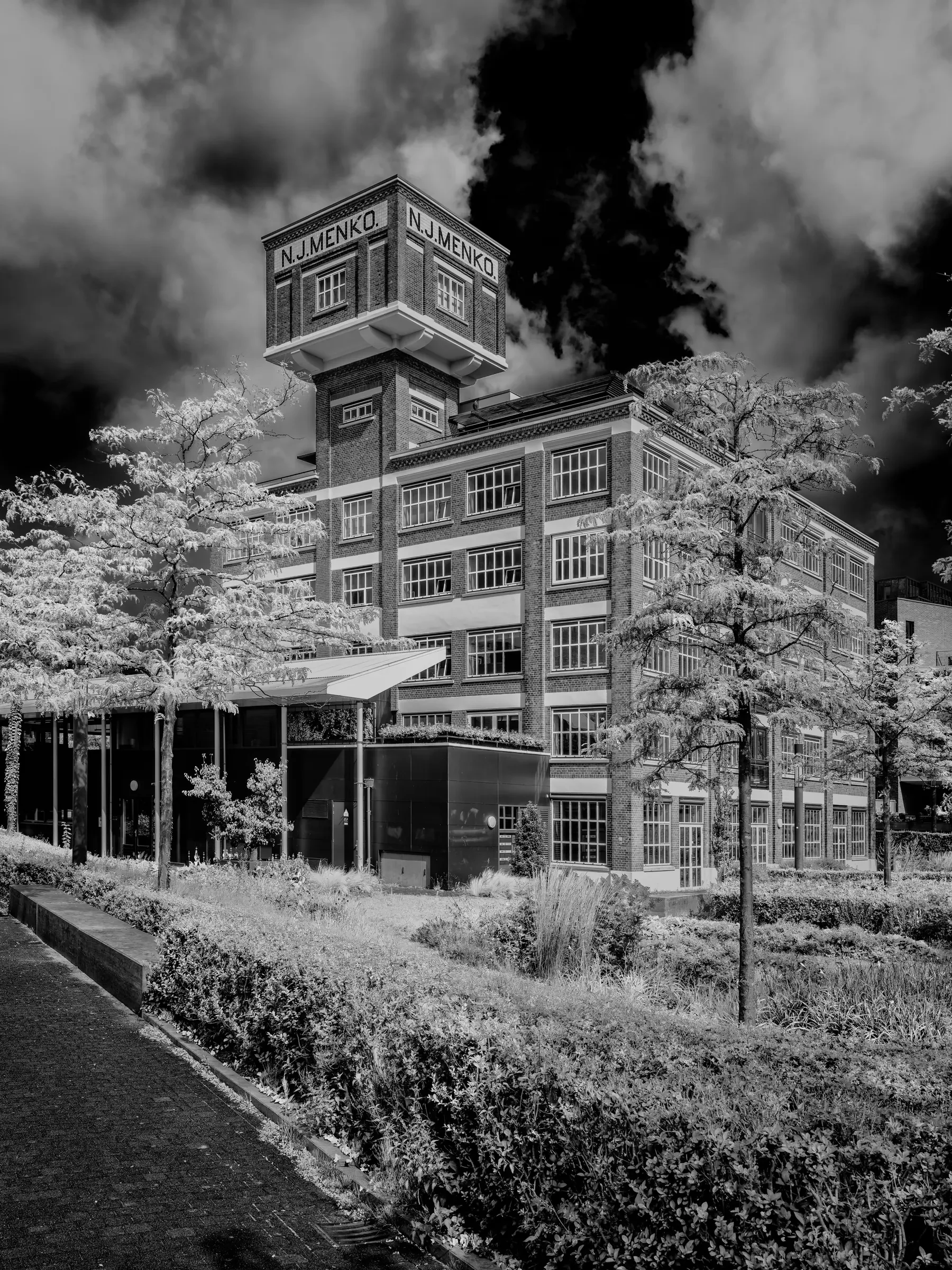 Black and white photograph of the N.J. Menko factory building in Enschede, with striking clouds and leafy trees in the foreground