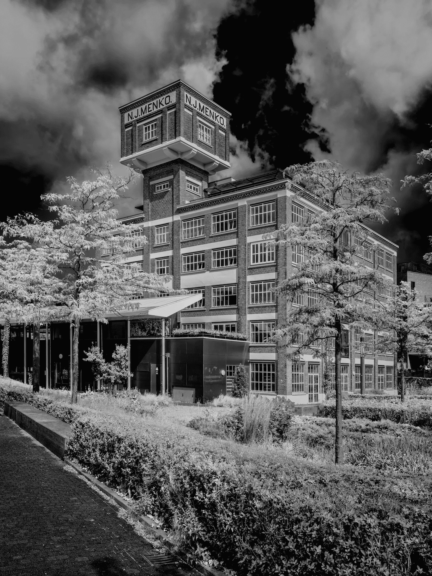 N.J. Menko Factory, Enschede Black and white photograph of the N.J. Menko factory building in Enschede, with striking clouds and leafy trees in the foreground